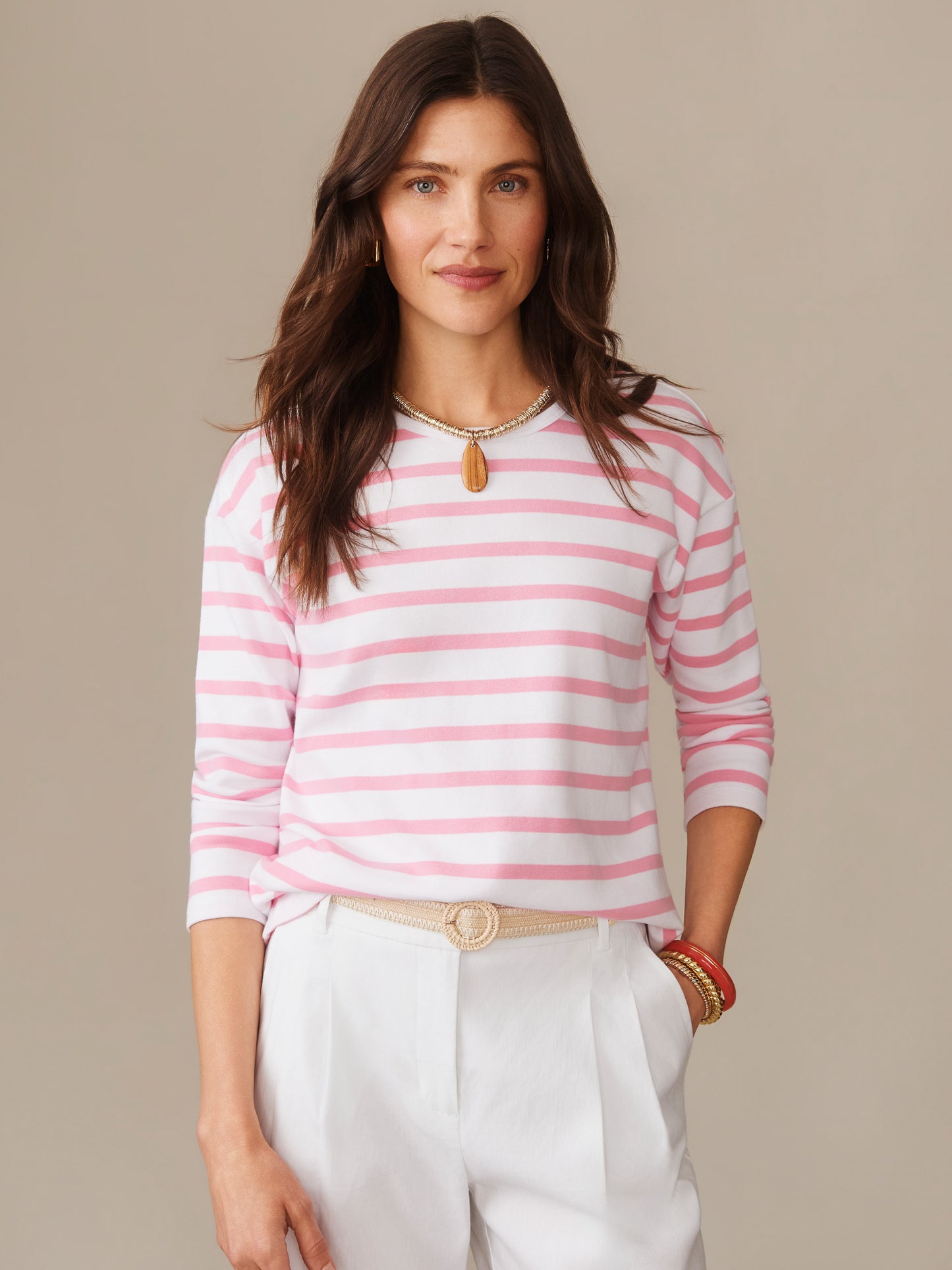Woman wearing a J. McLaughlin Nico Tee in Stripe with soft pink and white horizontal stripes, paired with white pants and accessorized with a necklace and bracelets, showcasing effortlessly stylish, enduring designs and innovative fabrics from a destination for defining style. Life, Well-Lived.