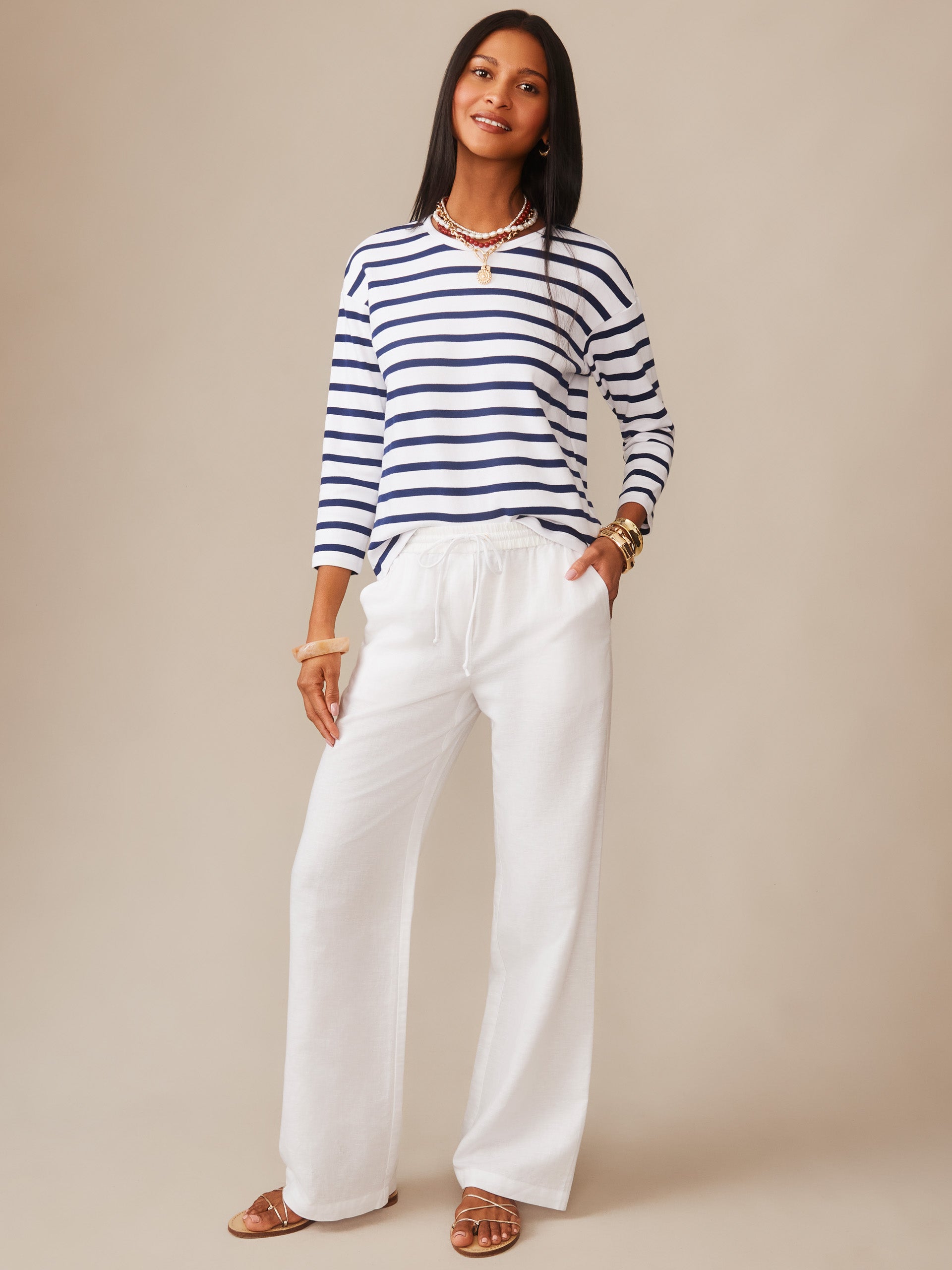 Woman wearing J.McLaughlin Nico Tee in Stripe with navy and white stripes, paired with white pants and gold accessories, showcasing effortlessly stylish, enduring designs and innovative fabrics.
