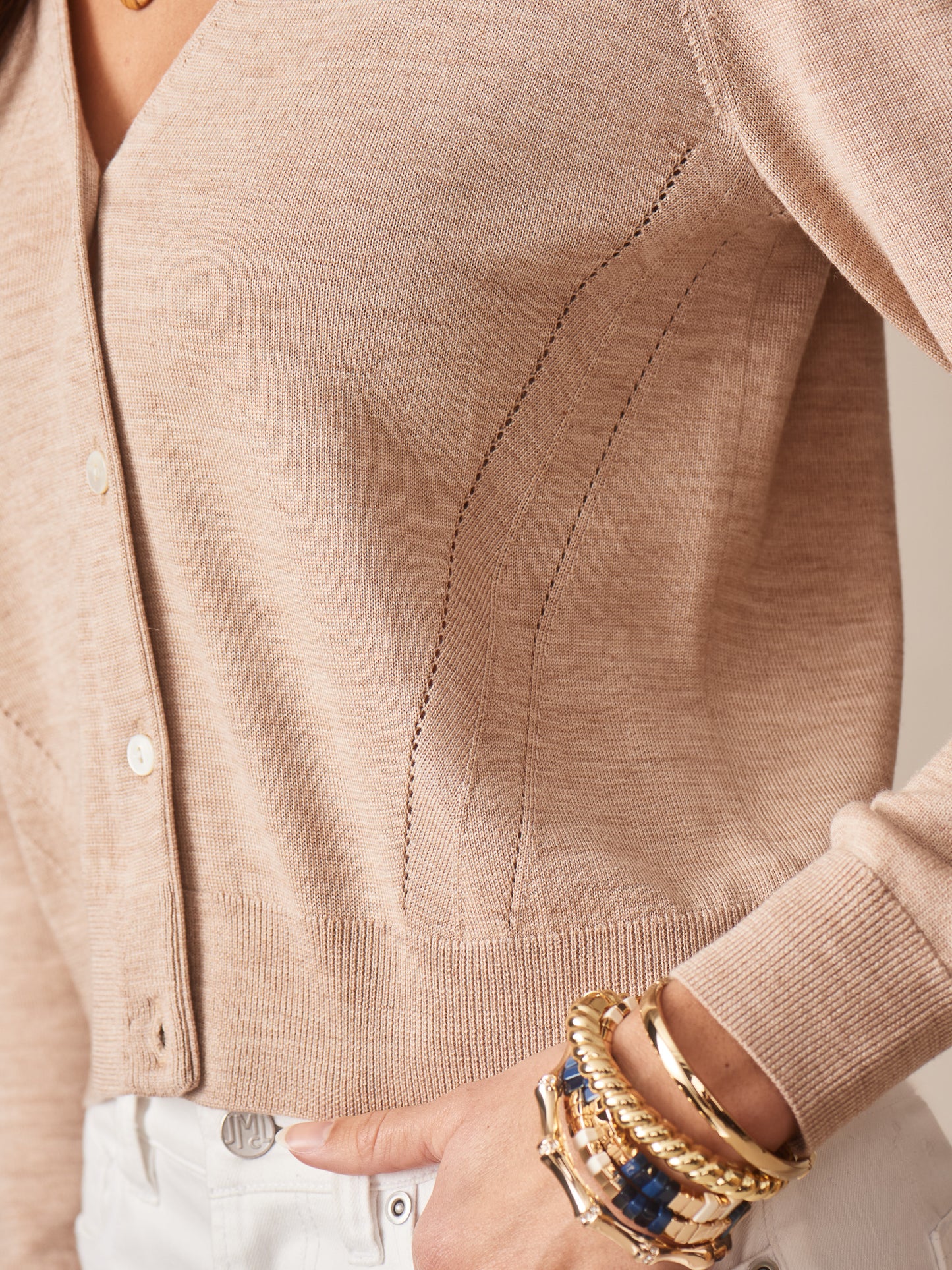 Close-up of a beige Olivette Cardigan by J.McLaughlin with delicate knit detailing paired with white pants and gold and blue bracelets, showcasing effortlessly stylish, enduring designs and innovative fabrics.