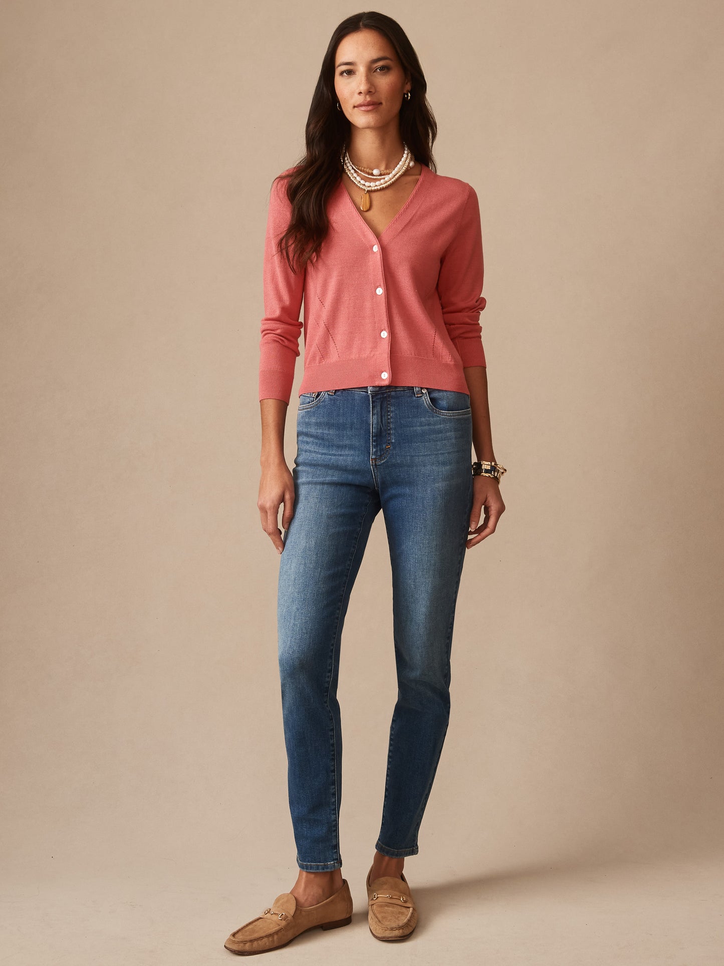 Woman wearing a soft coral Olivette Cardigan by J.McLaughlin paired with blue jeans and layered pearl necklaces, showcasing a timeless, classic sweater style.