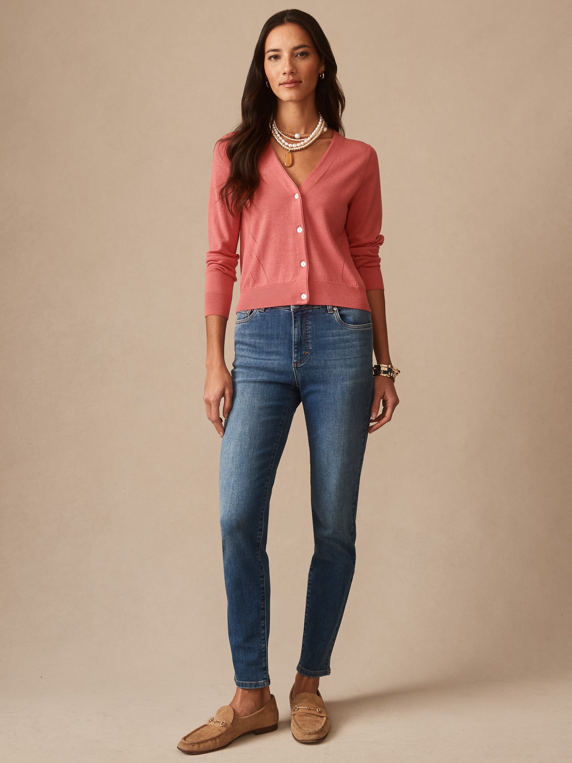 Woman wearing a soft coral Olivette Cardigan by J.McLaughlin paired with blue jeans and layered pearl necklaces, showcasing a timeless, classic sweater style.