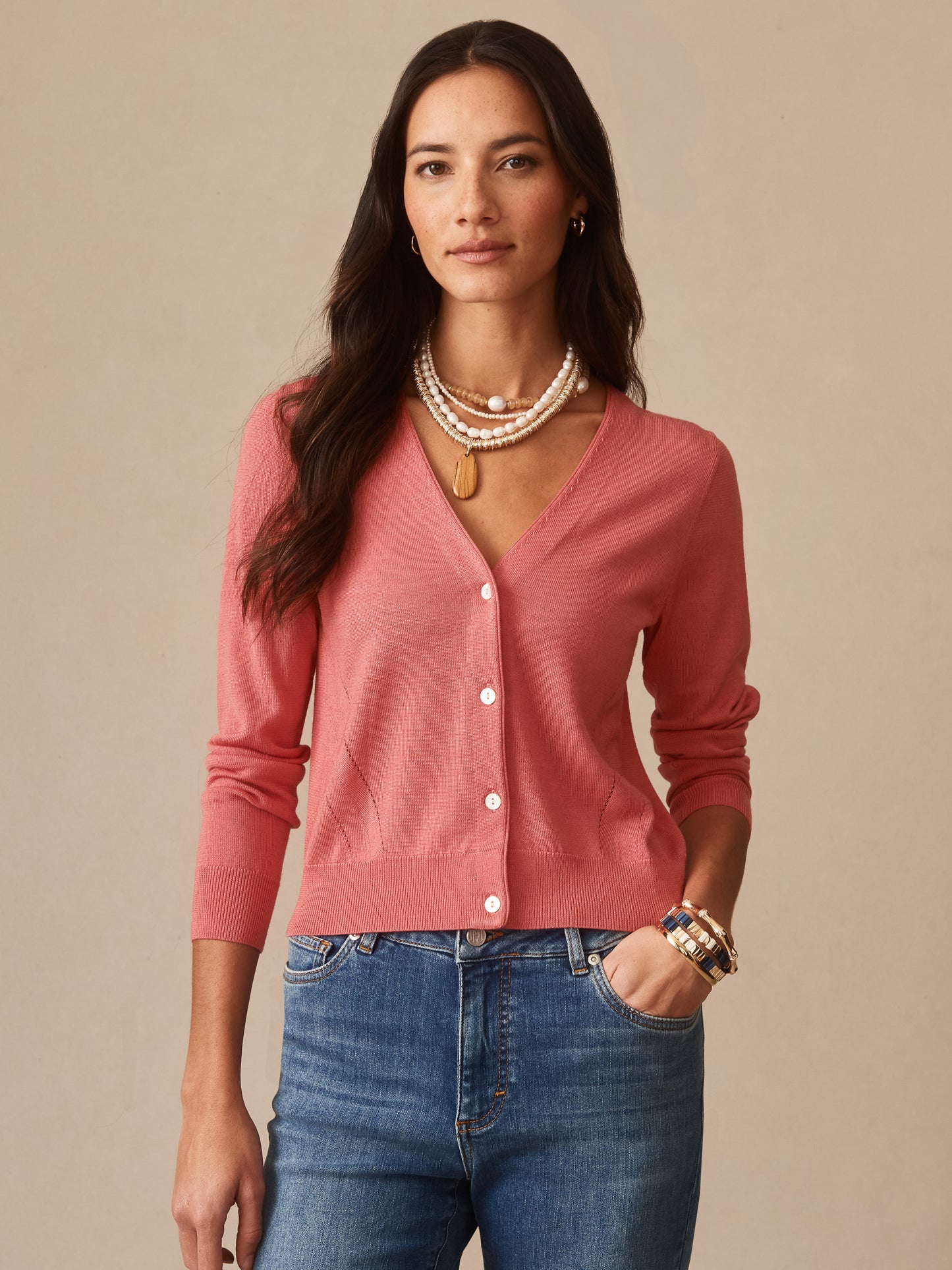 Woman wearing a soft coral Olivette Cardigan by J.McLaughlin, paired with blue jeans and layered pearl necklaces, showcasing timeless, classic style.