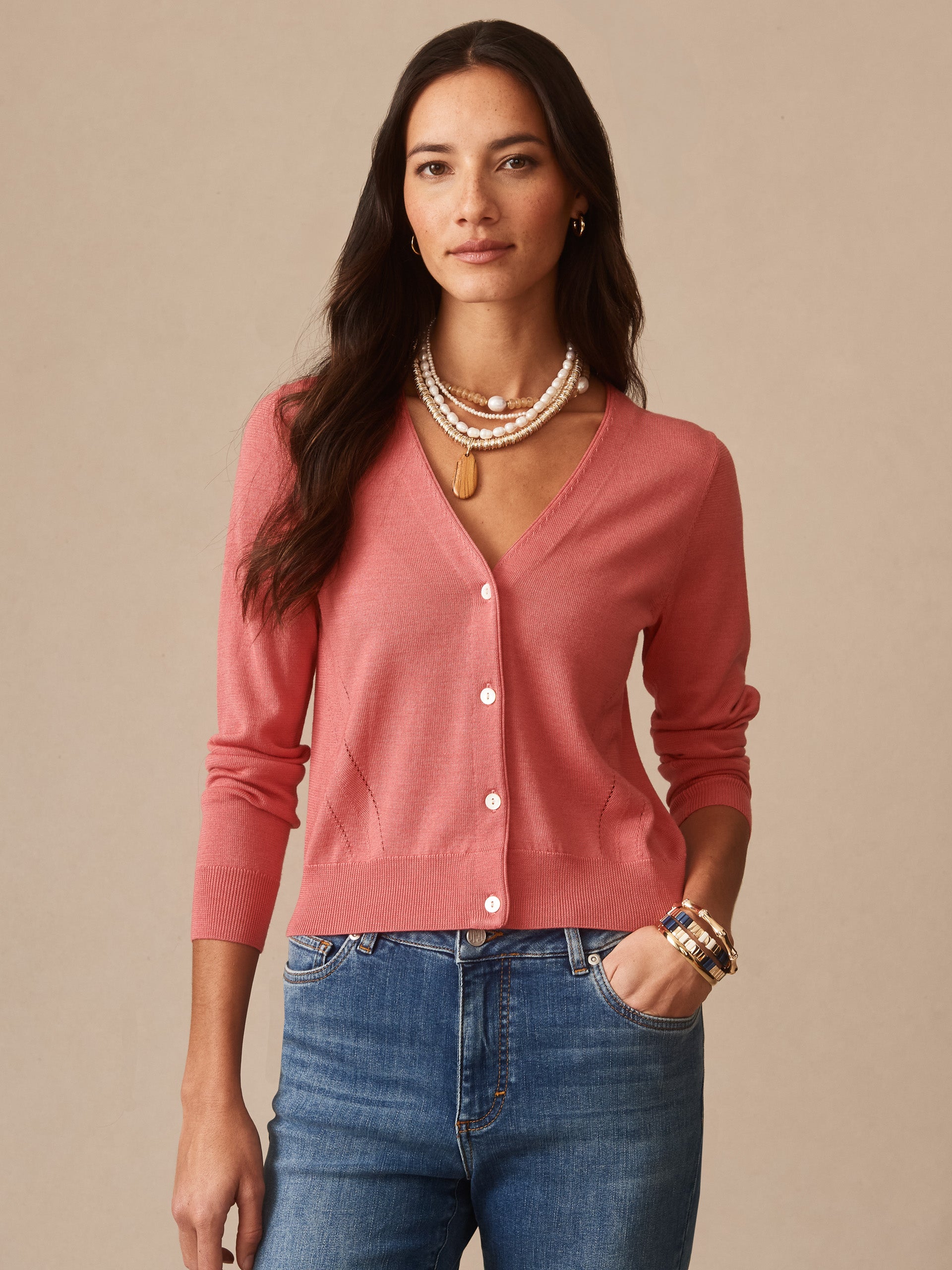 Woman wearing a soft coral Olivette Cardigan by J.McLaughlin, paired with blue jeans and layered pearl necklaces, showcasing timeless, classic style.