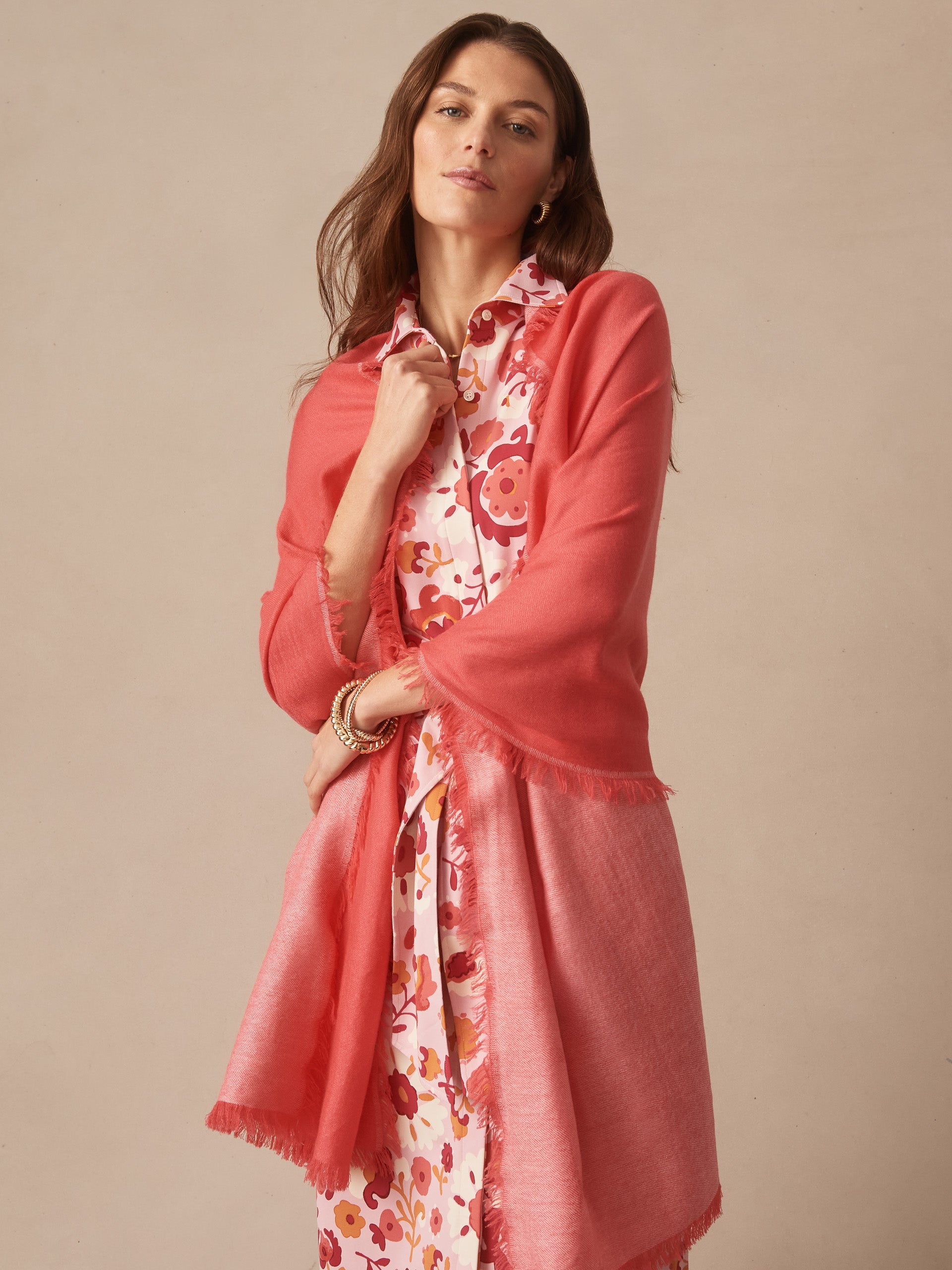 A woman wearing a coral pink Carey Cashmere Scarf by J.McLaughlin over a floral dress, embodying effortlessly stylish and enduring designs in innovative fabrics.