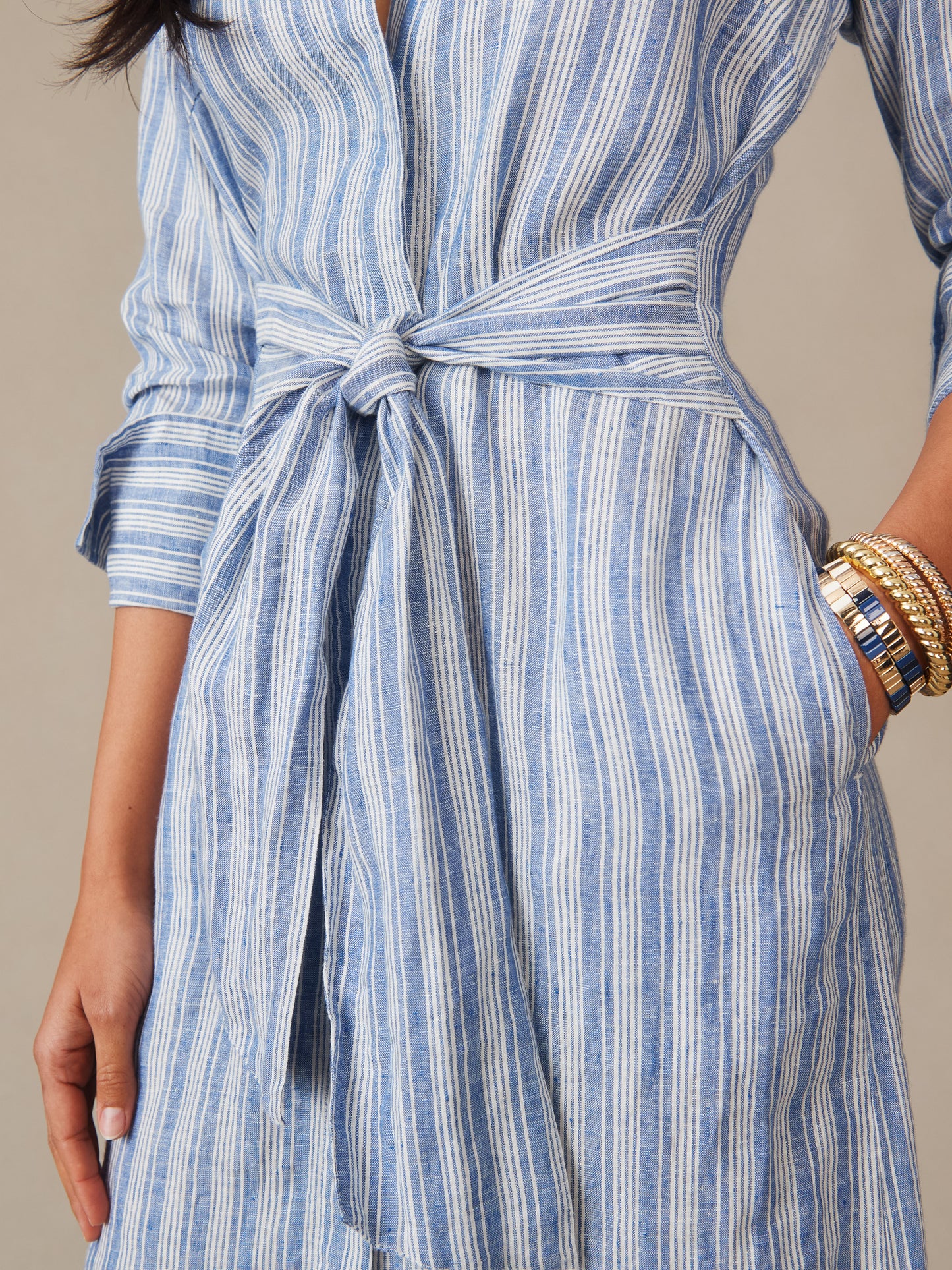 Close-up of a woman wearing the Orla Linen Dress in Stripe by J.McLaughlin, featuring a blue and white striped pattern, tied waist belt, and gold bracelets, embodying effortlessly stylish, enduring designs with innovative fabrics in a legacy of timeless fashion. A destination for defining style. Life, Well-Lived.
