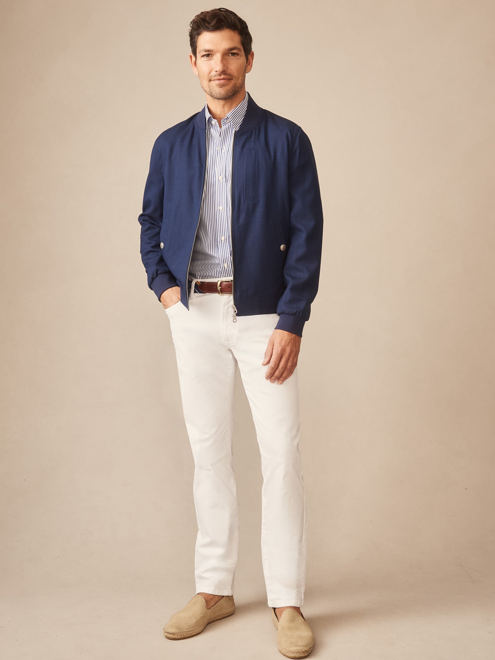 A man wearing the Outfield Jacket by J.McLaughlin in navy, paired with white pants and a striped shirt, showcasing effortlessly stylish, enduring designs and innovative fabrics from a legacy of timeless fashion. A destination for defining style. Life, Well-Lived.