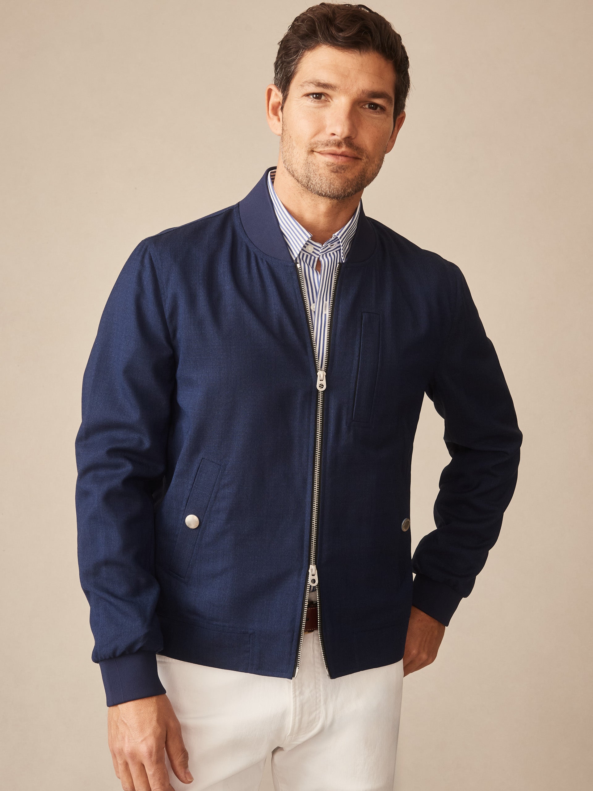 A man wearing a navy Outfield Jacket by J.McLaughlin, styled with a striped shirt and white pants, showcasing effortlessly stylish, enduring designs and innovative fabrics.
