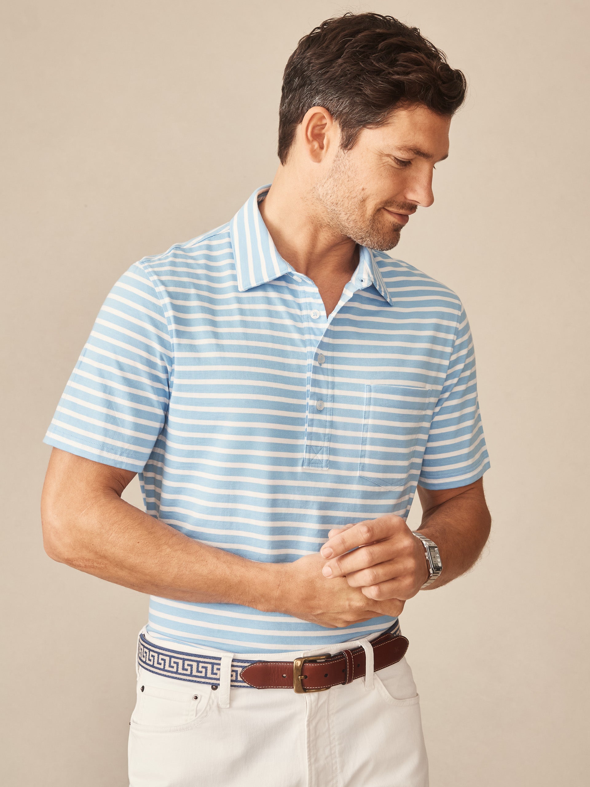 A man wears the Owens Polo in Breton Stripe by J.McLaughlin, featuring soft blue and white stripes, paired with white pants and a stylish belt, showcasing effortlessly stylish, enduring designs in a legacy of timeless fashion. A destination for defining style. Life, Well-Lived.