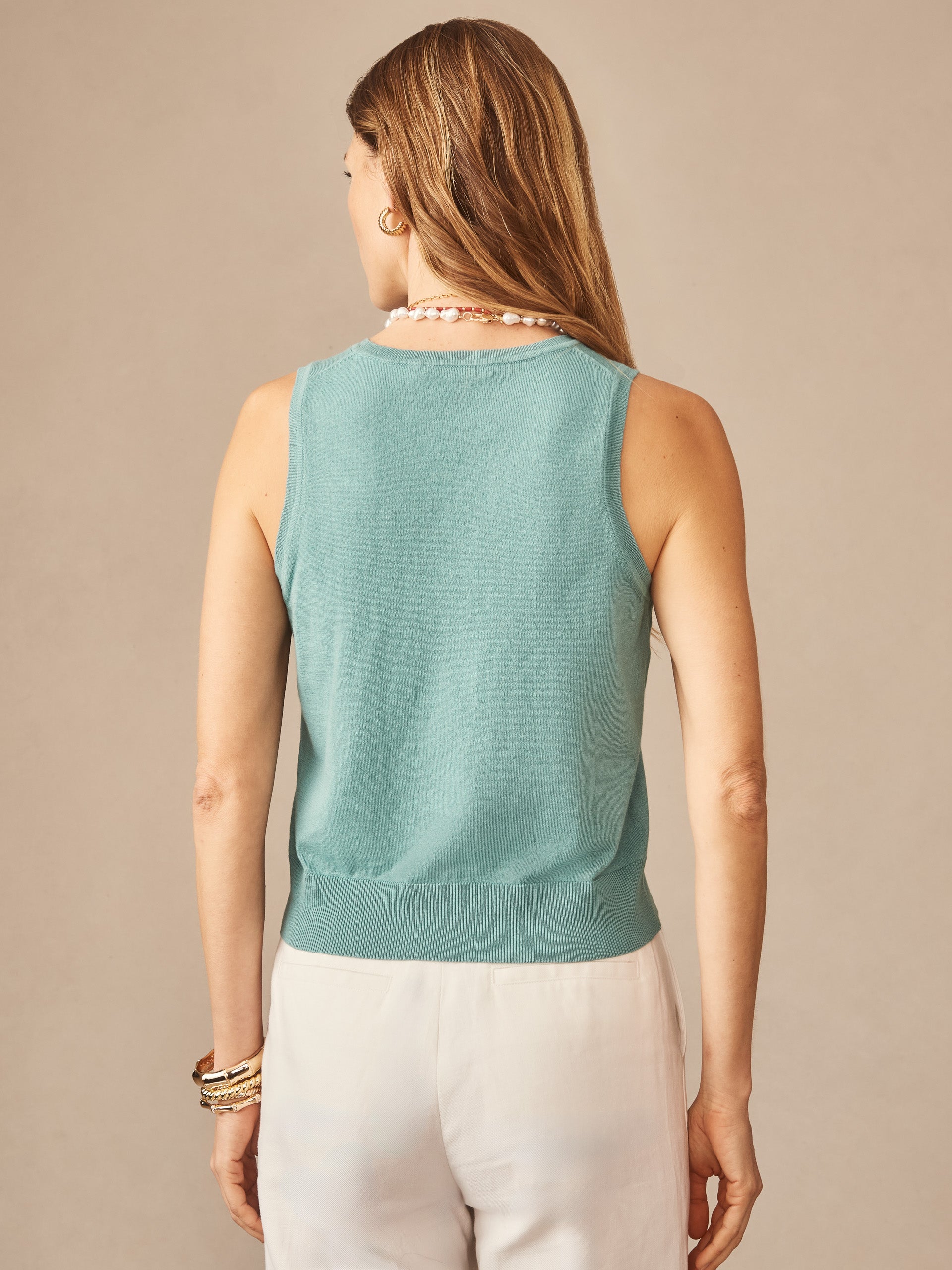 Woman wearing a turquoise Opal Sweater Tank by J.McLaughlin, styled with white pants and accessorized with bracelets and a necklace, showcasing effortlessly stylish and enduring designs.