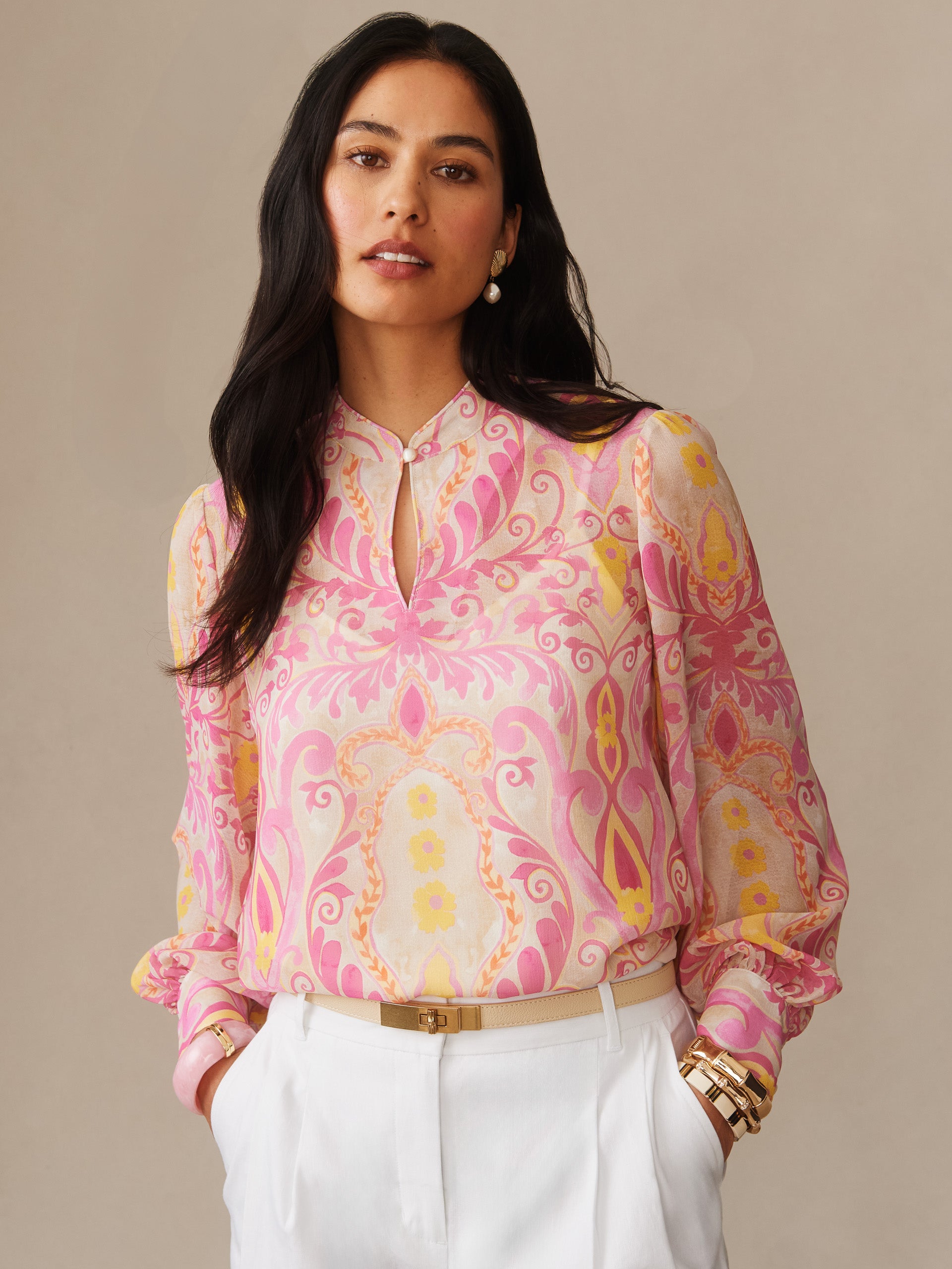 Woman wearing Peyton Blouse in Mini Deco Bloom by J.McLaughlin featuring soft pink and cream floral patterns, styled with white pants and gold accessories, showcasing effortlessly stylish, enduring designs and a legacy of timeless fashion. A destination for defining style. Life, Well-Lived.