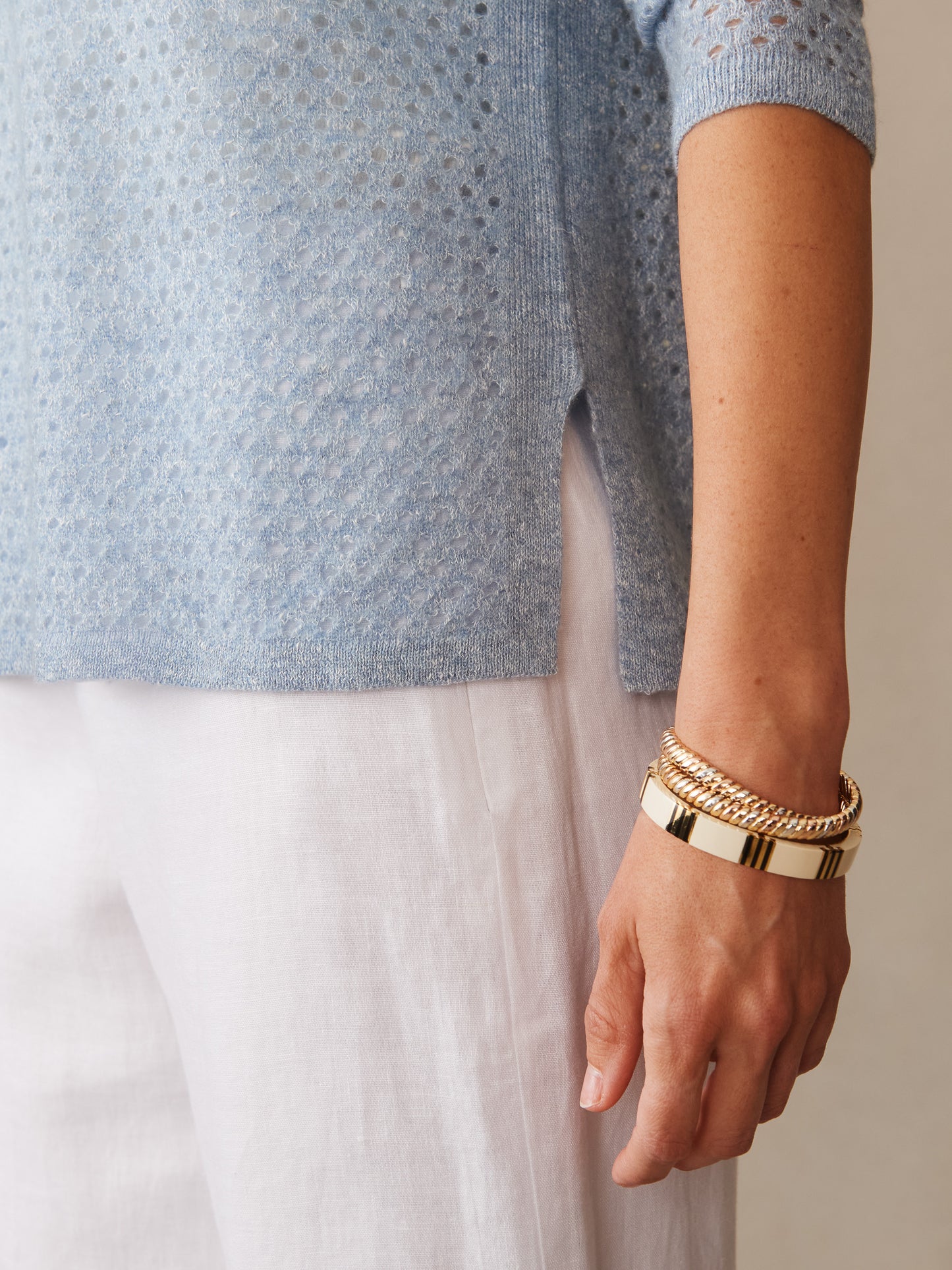 Close-up of a light blue Philomena Cashmere Linen Sweater from J.McLaughlin paired with white linen pants and gold-tone bracelets, showcasing effortlessly stylish, enduring designs with innovative fabrics.