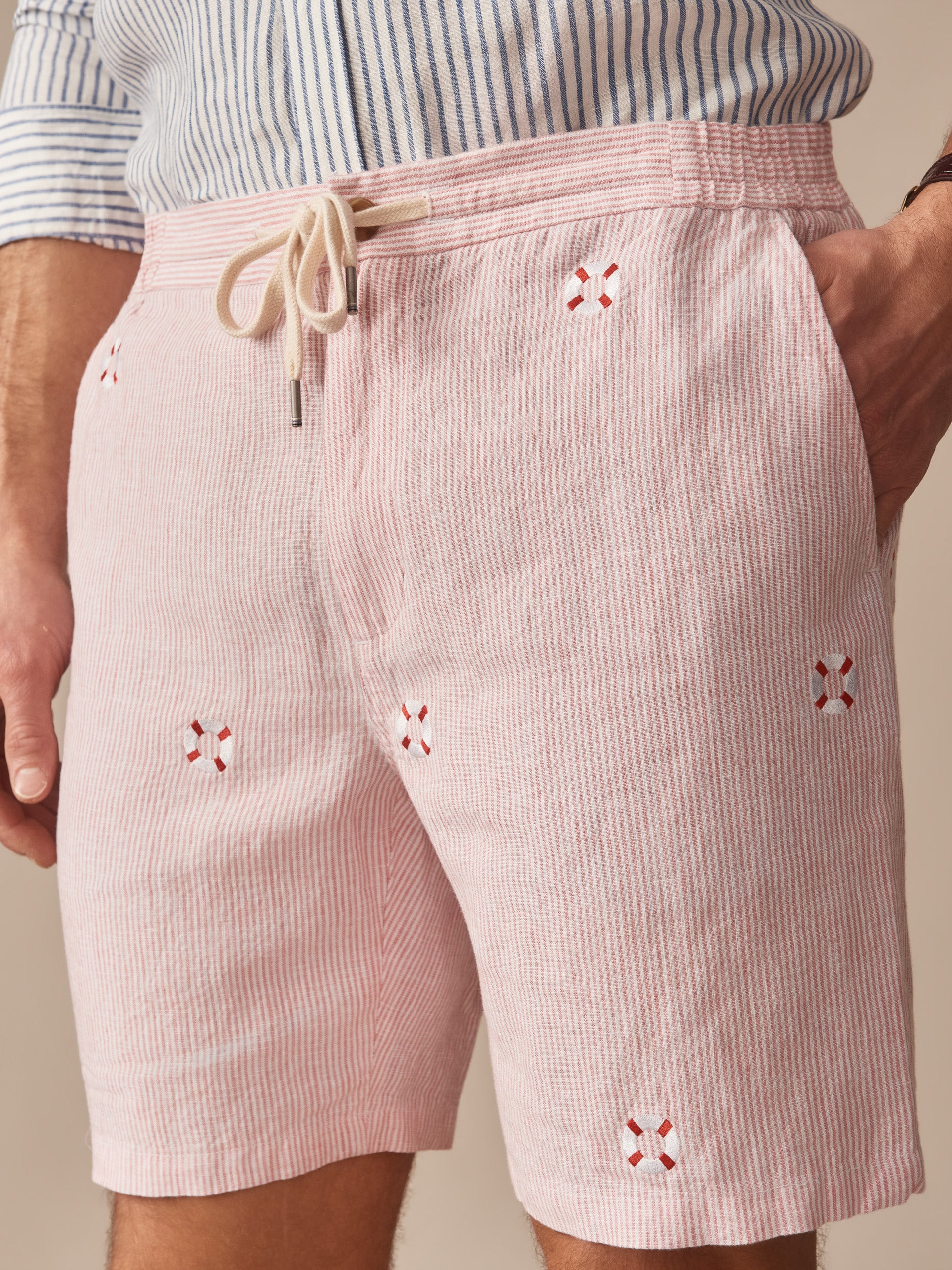 A close-up of Newport Linen Shorts in Lifesaver by J.McLaughlin featuring a classic pink striped pattern with embroidered detailing and a drawstring waist, showcasing effortlessly stylish design with enduring fabrics from a brand known for timeless fashion. A destination for defining style. Life, Well-Lived.