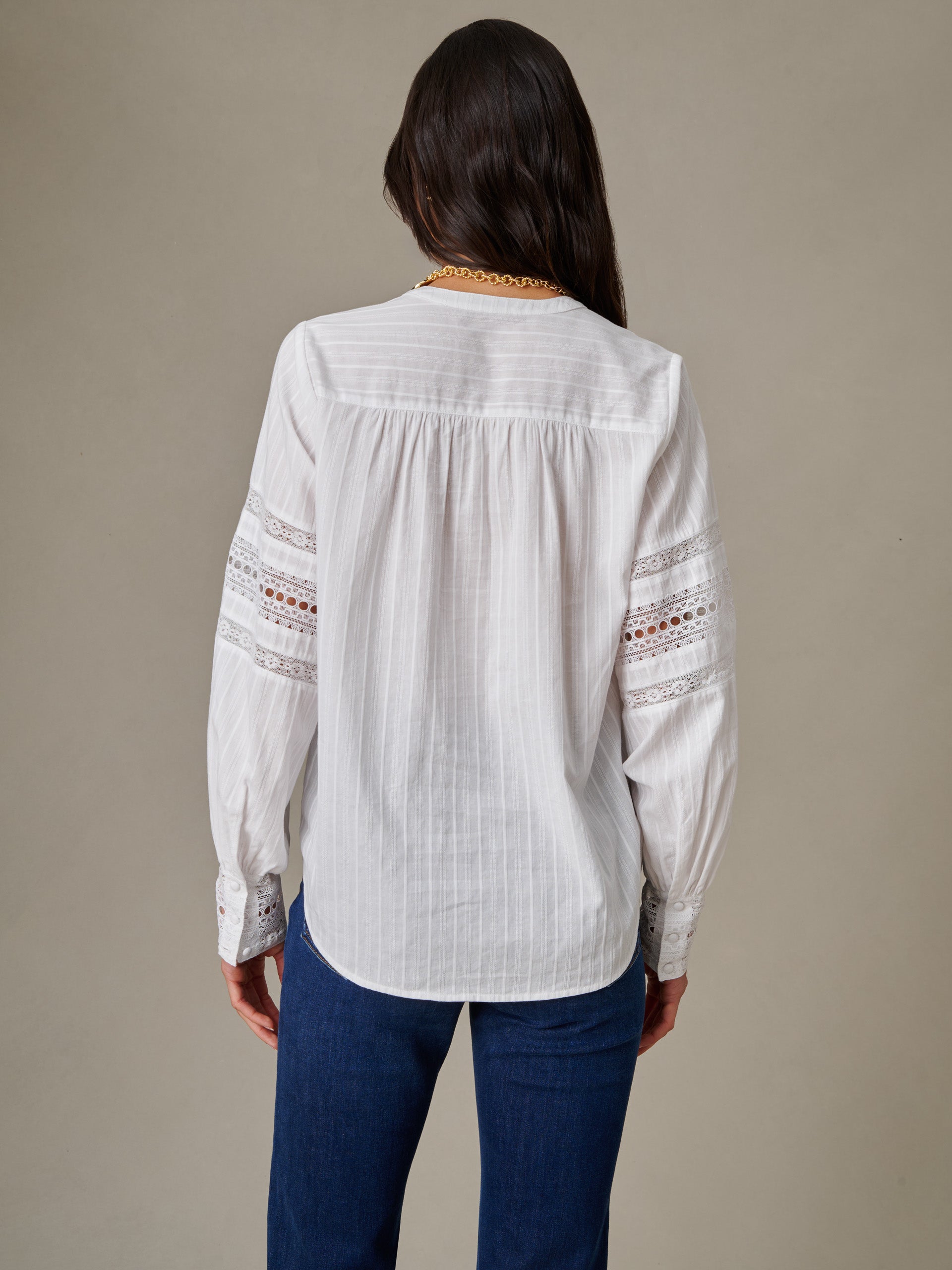 Back view of a woman wearing a white Posey Blouse by J.McLaughlin, featuring delicate lace inserts on the sleeves, paired with dark blue jeans in a timeless, effortless style.