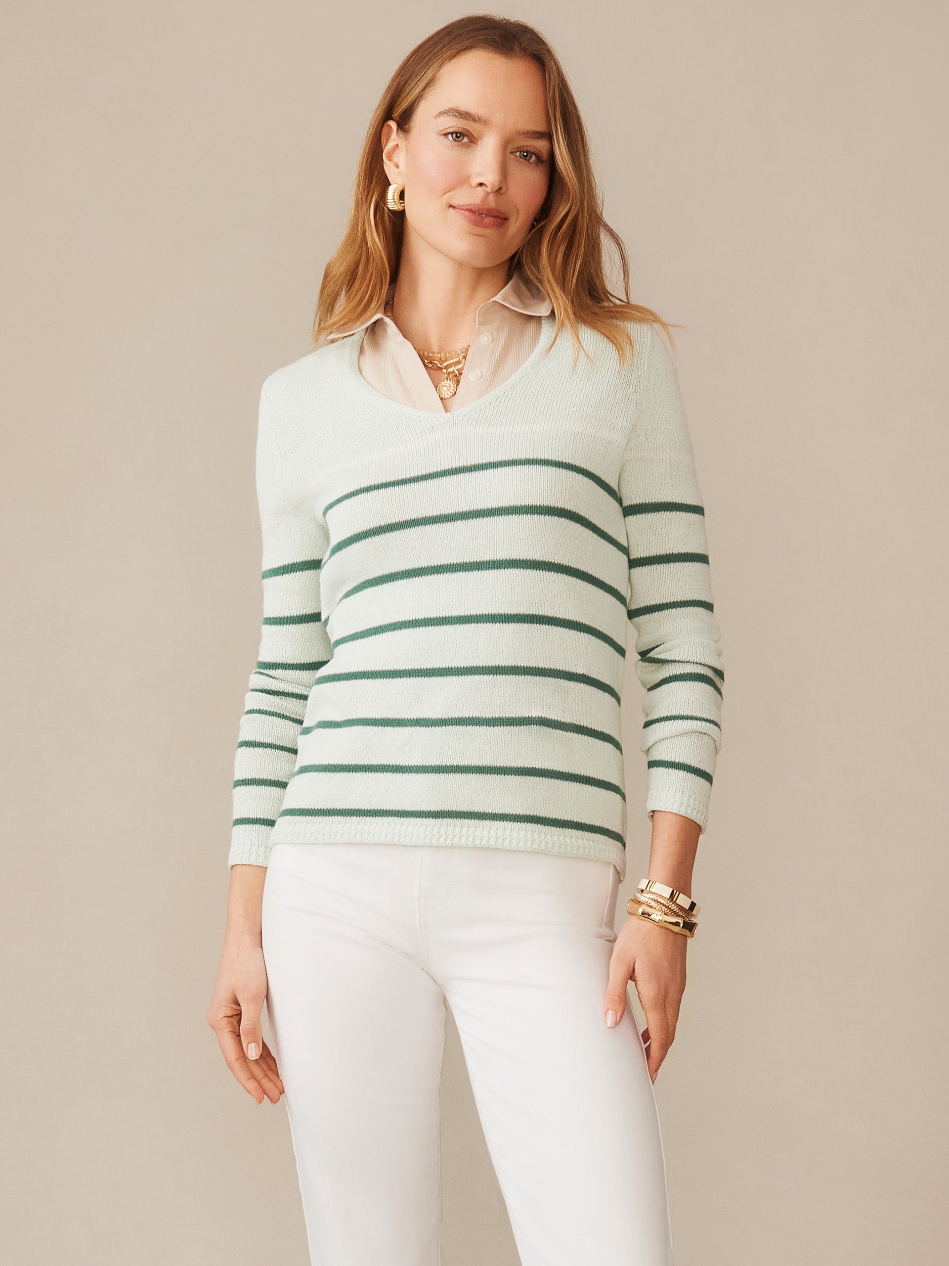 A woman models the Ramona Linen Blend Sweater in Stripe by J.McLaughlin, featuring green stripes on a light background, paired with white pants and gold accessories, showcasing effortlessly stylish, enduring designs with innovative fabrics in a legacy of timeless fashion. A destination for defining style. Life, Well-Lived.