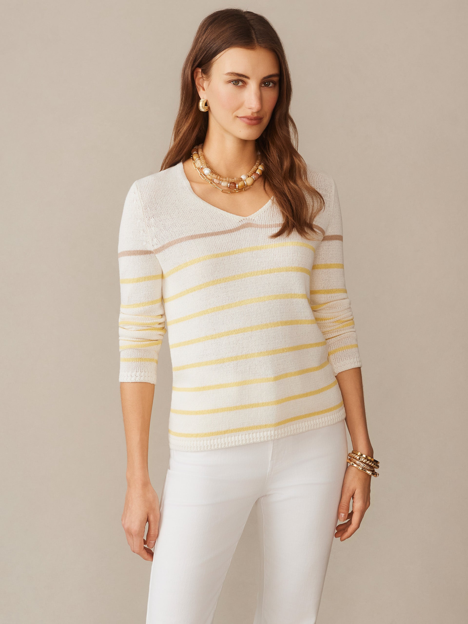 A woman wearing the Ramona Linen Blend Sweater in Stripe by J.McLaughlin, styled effortlessly with white pants and gold accessories, showcasing enduring designs and innovative fabrics.