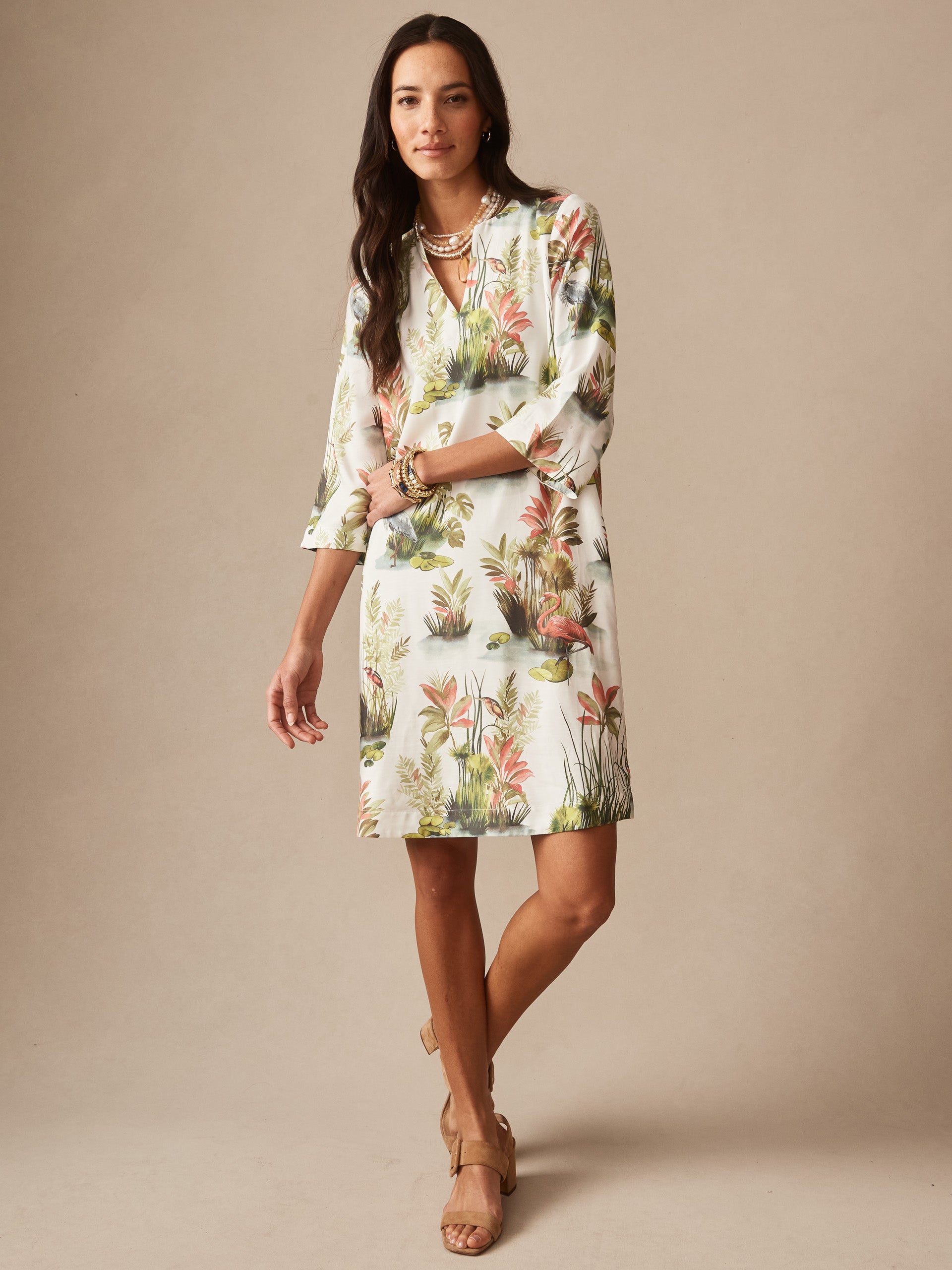 Woman wearing the Roni Dress in Tidal Palms by J.McLaughlin, featuring a soft white fabric with coral and green tropical palm and flamingo print, styled with layered necklaces and bracelets, showcasing effortlessly stylish, enduring designs and innovative fabrics. A destination for defining style. Life, Well-Lived.