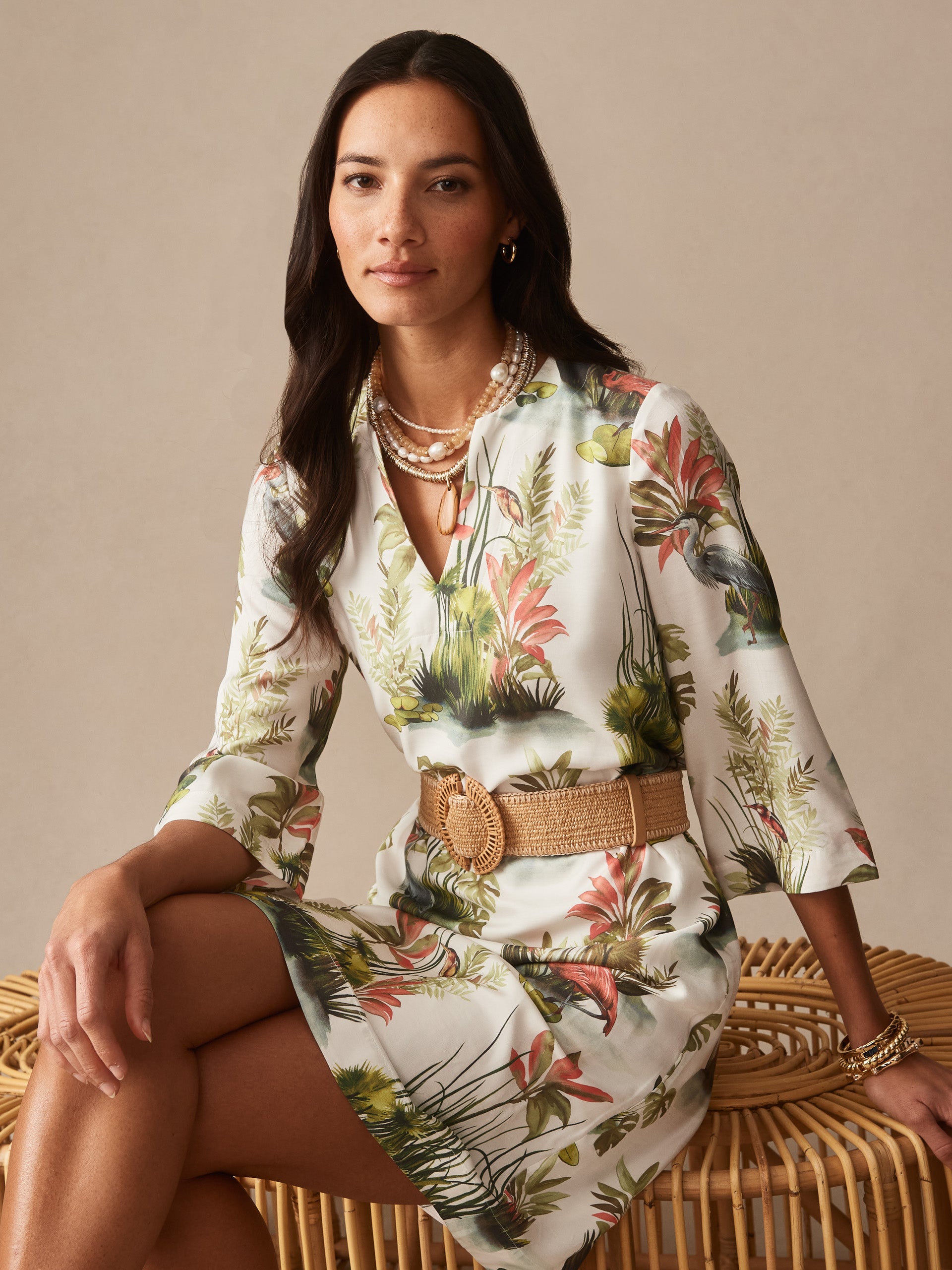 A woman wearing the Roni Dress in Tidal Palms by J.McLaughlin, featuring a tropical botanical print with coral, green, and white hues, styled with a woven belt and layered pearl necklaces, showcasing effortless style and enduring designs from a legacy of timeless fashion. A destination for defining style. Life, Well-Lived.