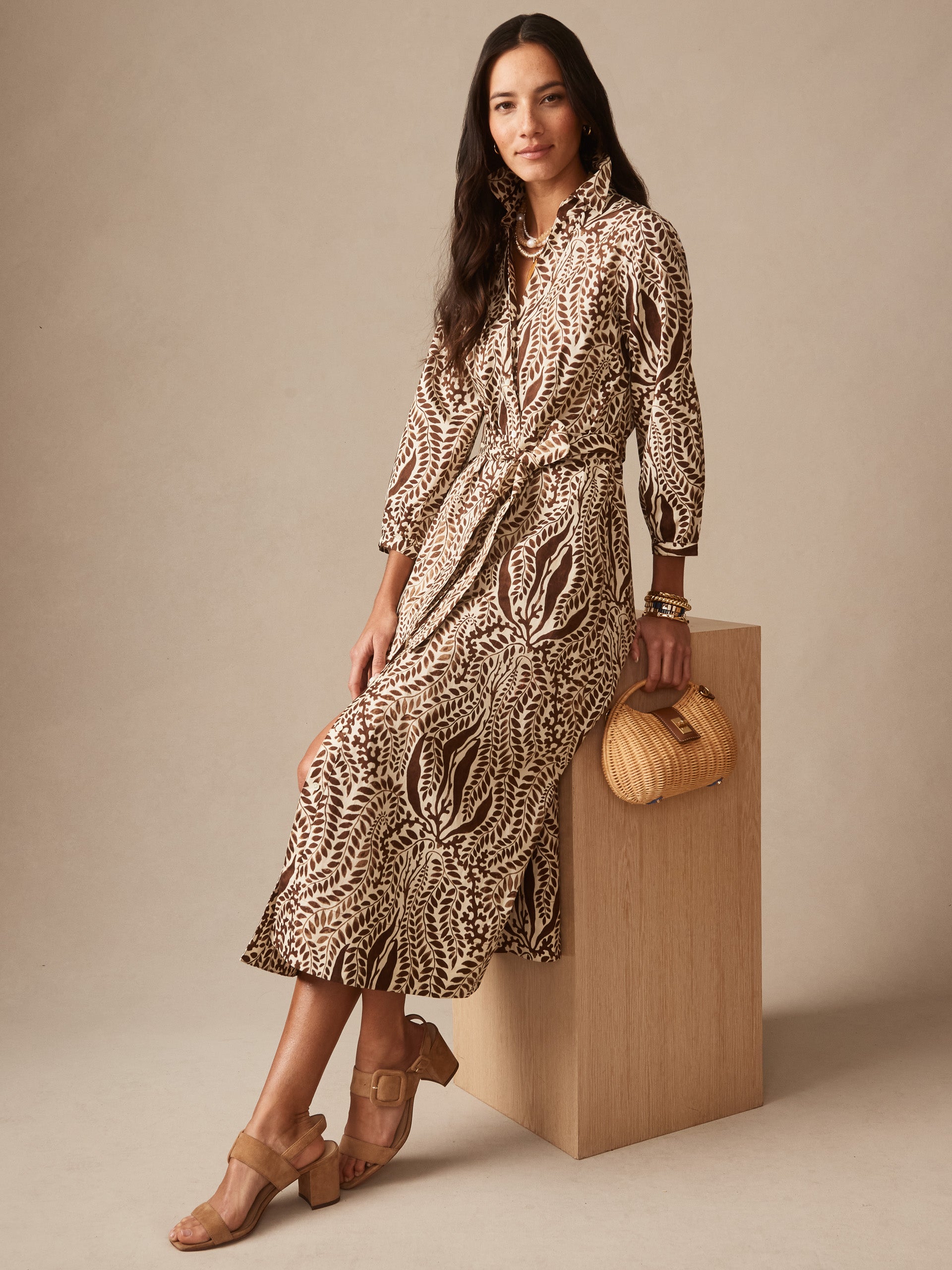 A woman wearing the Sena Dress in Coral Current by J.McLaughlin, featuring earthy leaf patterns, paired with tan heeled sandals and a woven handbag, showcasing effortlessly stylish, enduring designs with a legacy of timeless fashion. A destination for defining style. Life, Well-Lived.