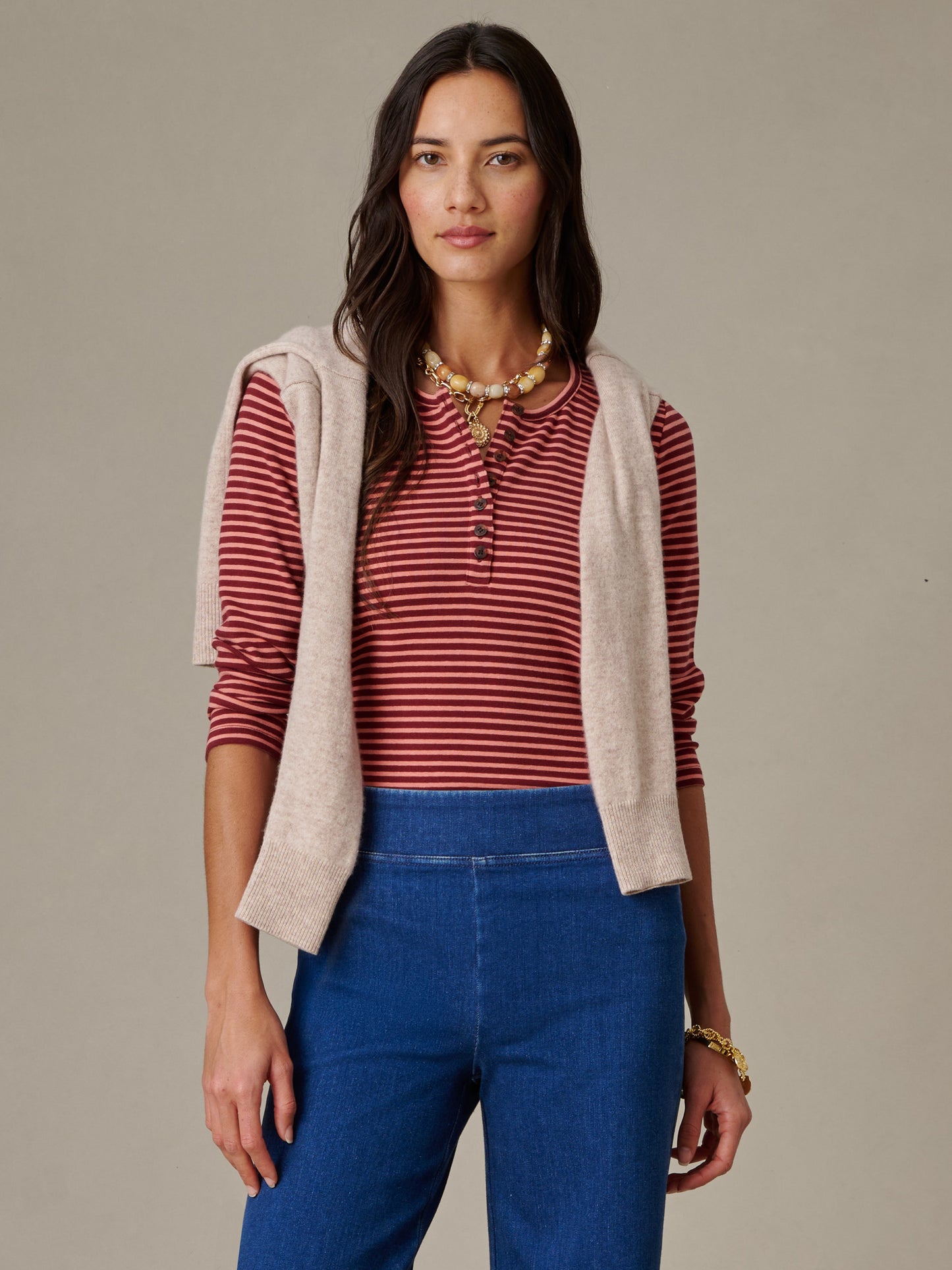 Woman wearing a Sinclair Henley in Stripe by J.McLaughlin paired with blue pants and a beige sweater draped over her shoulders, embodying effortlessly stylish, enduring designs.
