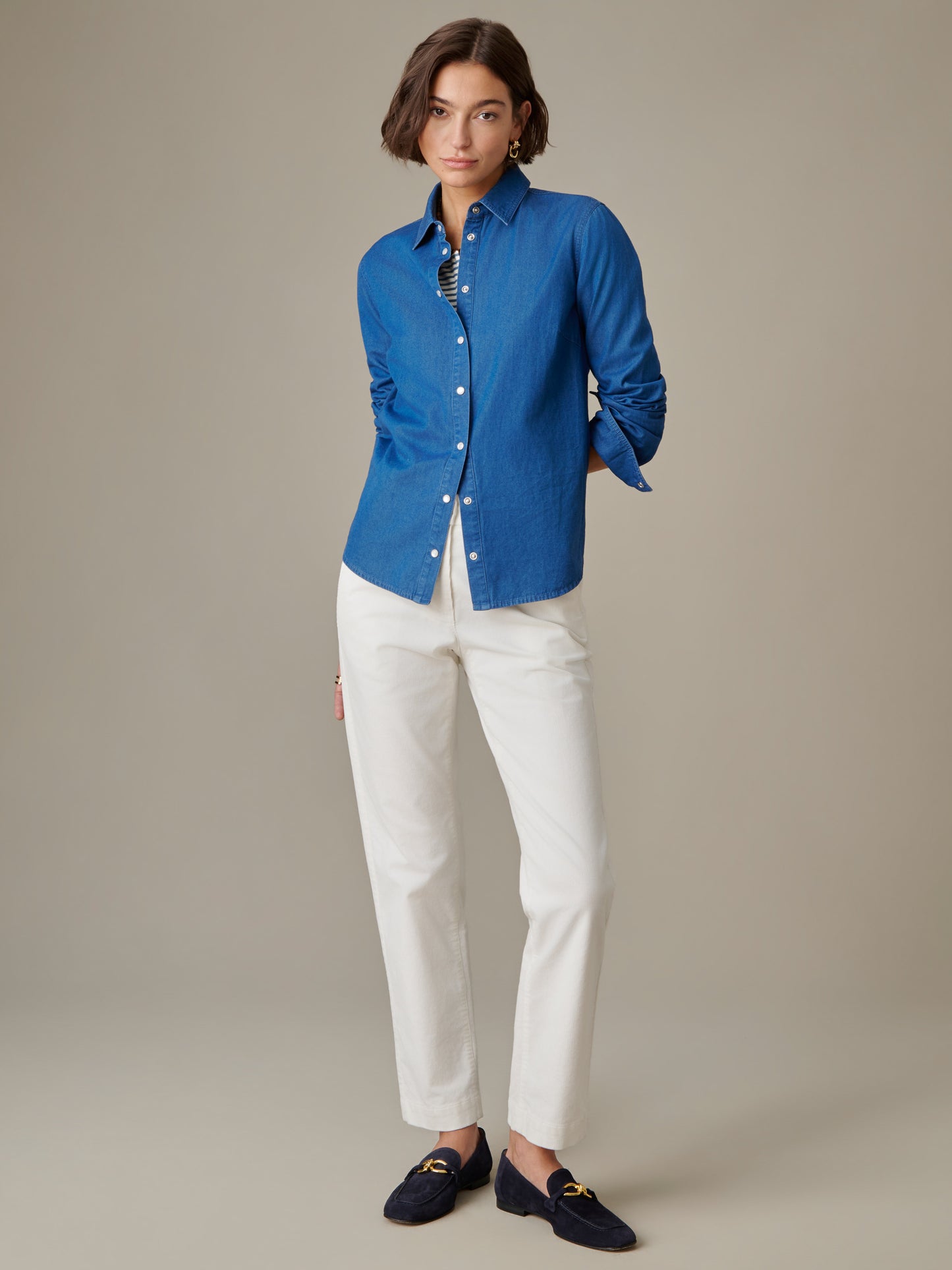 A woman wearing the Lois Chambray Shirt by J.McLaughlin in blue, styled with white pants and navy loafers, showcasing effortlessly stylish and enduring design.