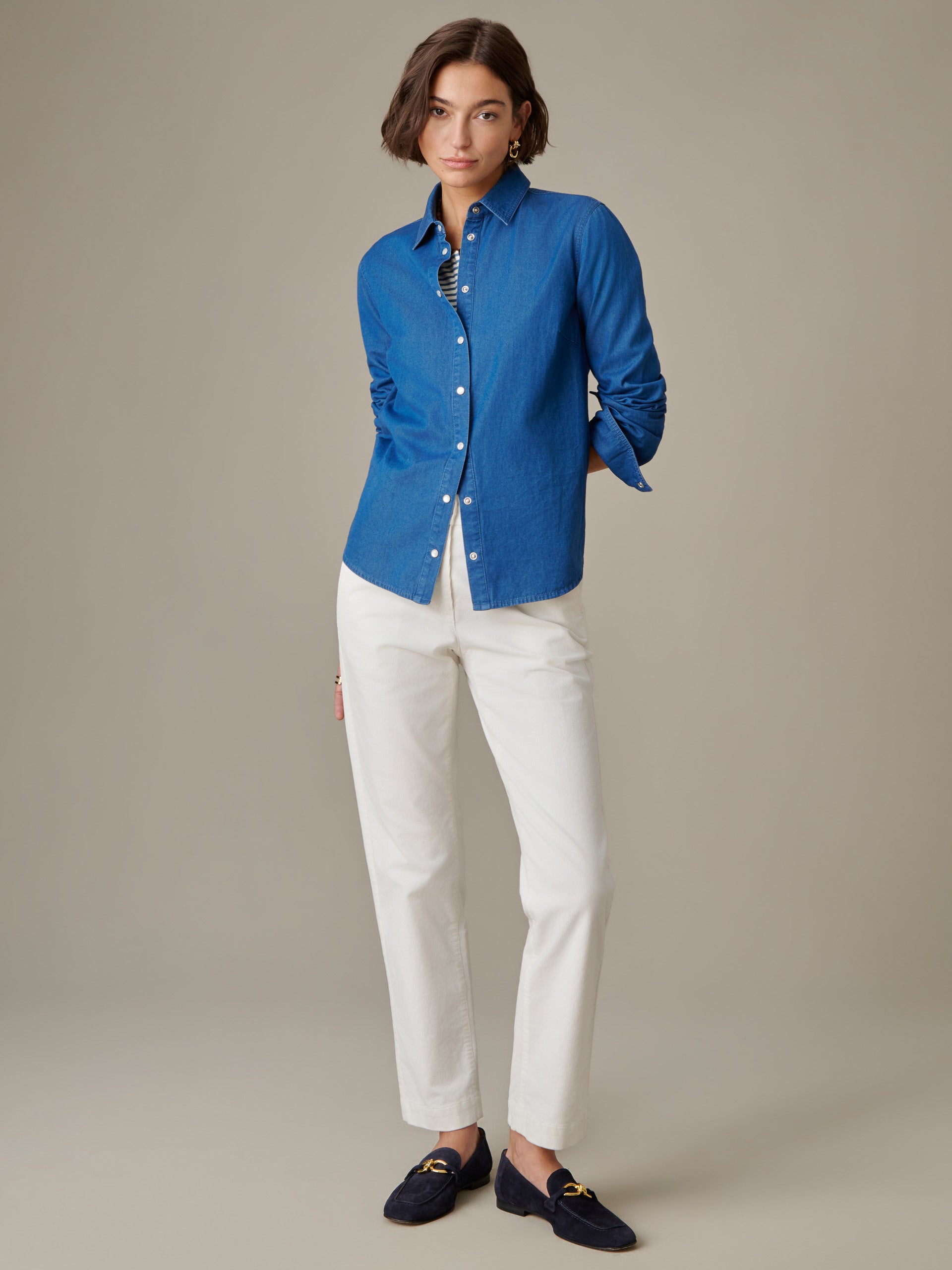 A woman wearing the Lois Chambray Shirt by J.McLaughlin in blue, styled with white pants and navy loafers, showcasing effortlessly stylish and enduring design.