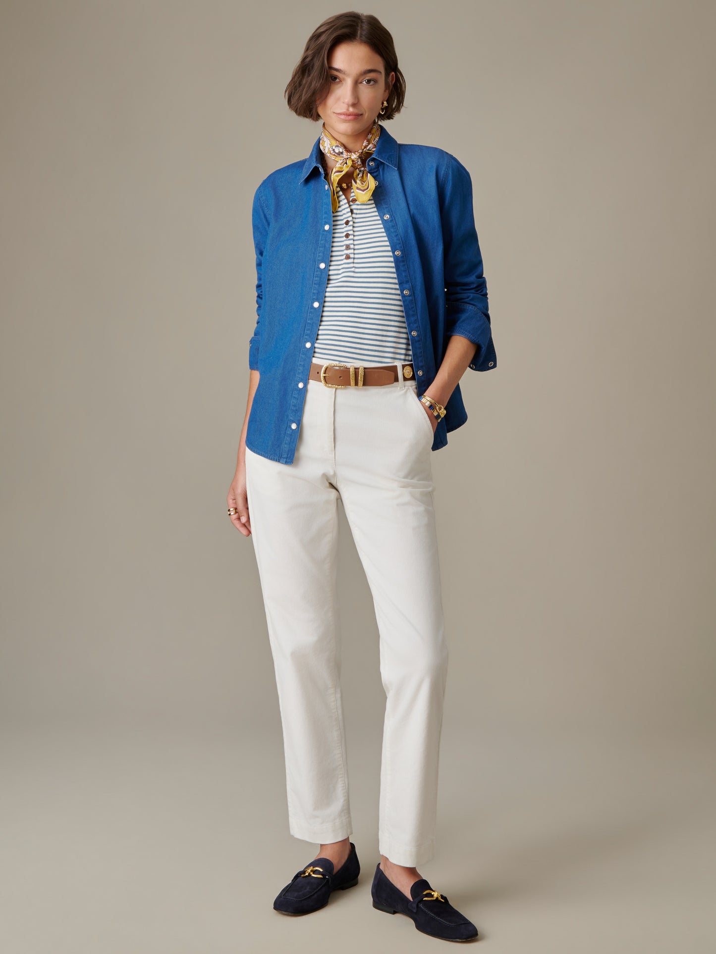 Woman wearing a blue Lois Chambray Shirt by J.McLaughlin over a striped top with cream pants, brown belt, navy loafers, and a yellow patterned scarf, showcasing timeless, effortless style.