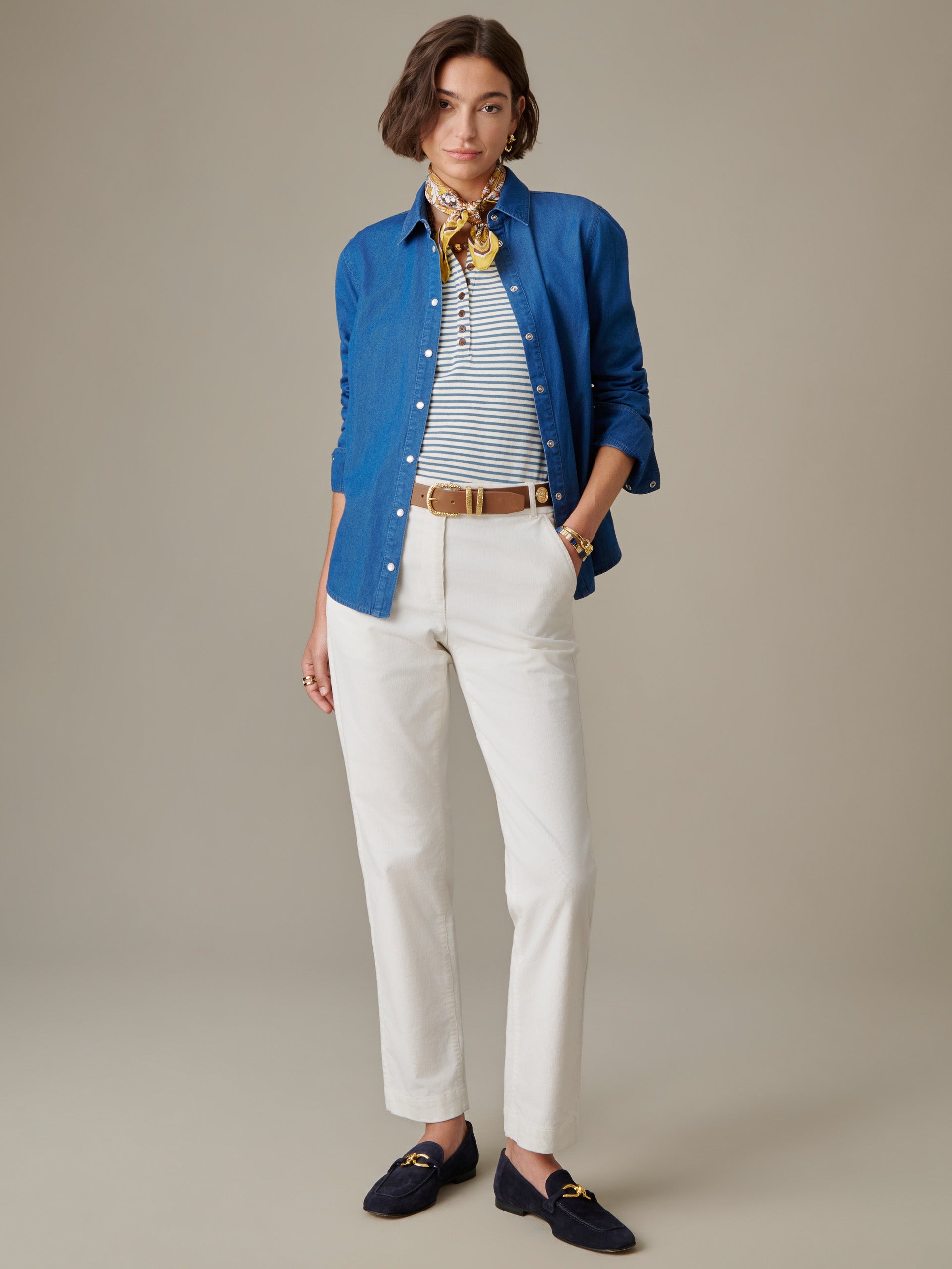Woman wearing a blue Lois Chambray Shirt by J.McLaughlin over a striped top with cream pants, brown belt, navy loafers, and a yellow patterned scarf, showcasing timeless, effortless style.