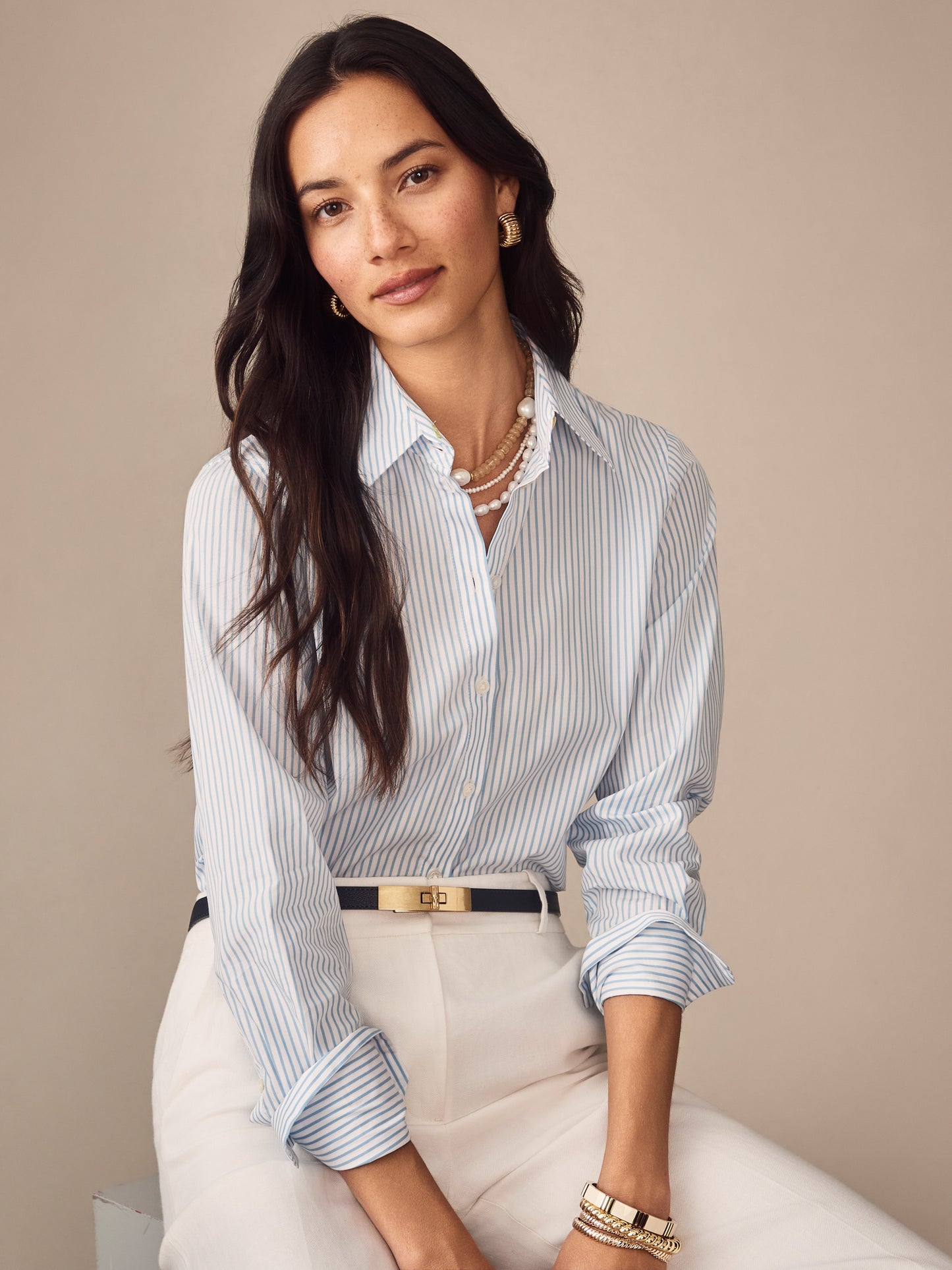 Woman wearing a J.McLaughlin Sloane Shirt in Stripe, featuring soft blue and white stripes, styled with white pants and elegant jewelry, showcasing effortlessly stylish, enduring designs.