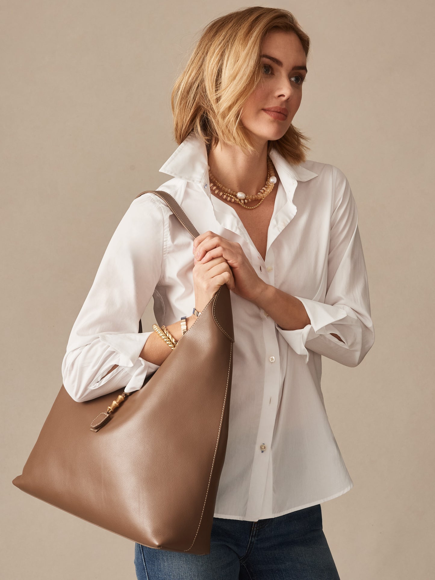 Woman wearing a white shirt and jeans carries a taupe Jenna Leather Tote by J.McLaughlin, showcasing effortlessly stylish and enduring designs in a classic accessory.