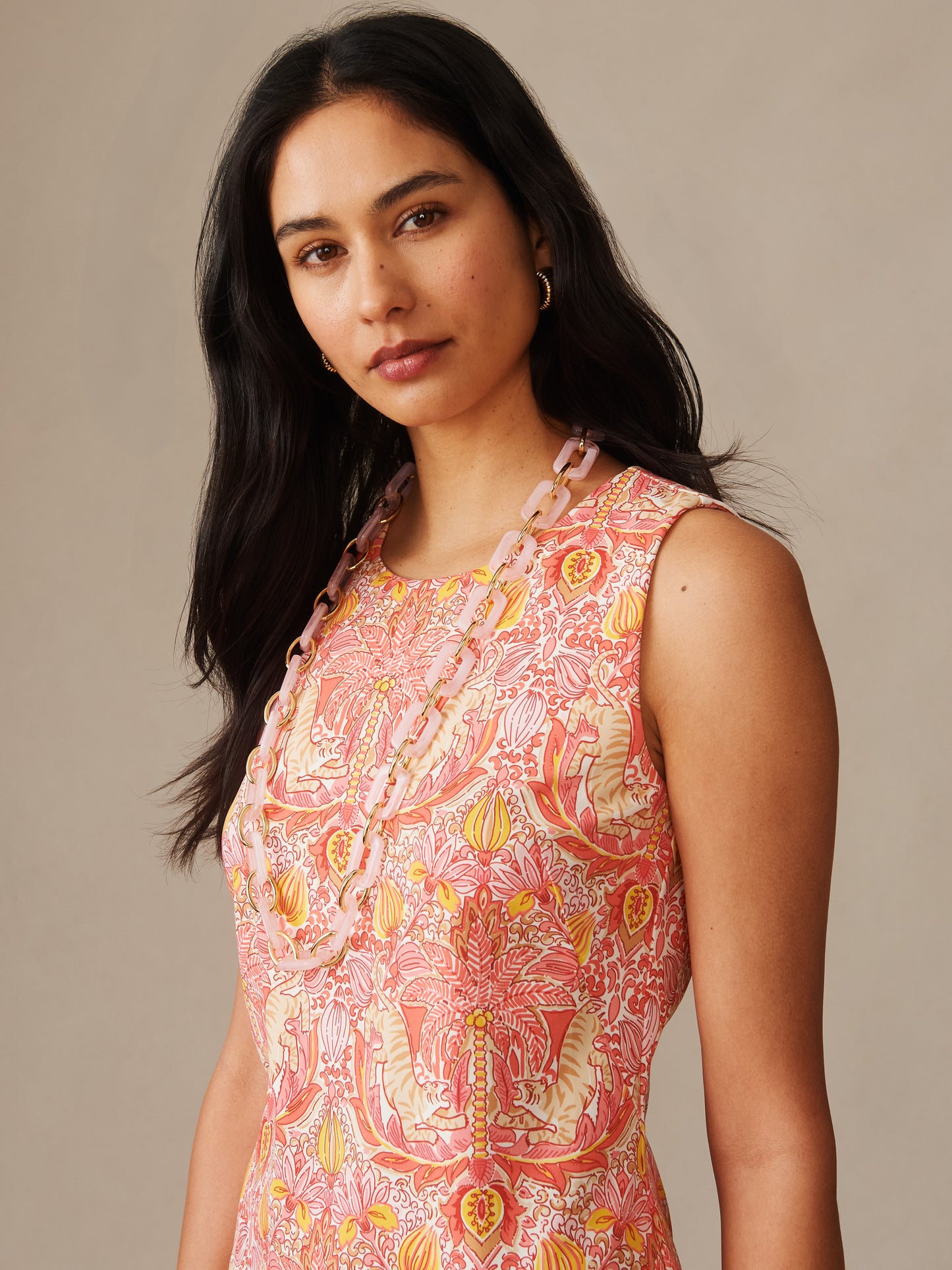 A model wearing the Sophia Sleeveless Dress in Tigress Tropico by J.McLaughlin, featuring an effortlessly stylish coral pink patterned dress with a statement necklace, embodying enduring designs and innovative fabrics from a destination for defining style. Life, Well-Lived.