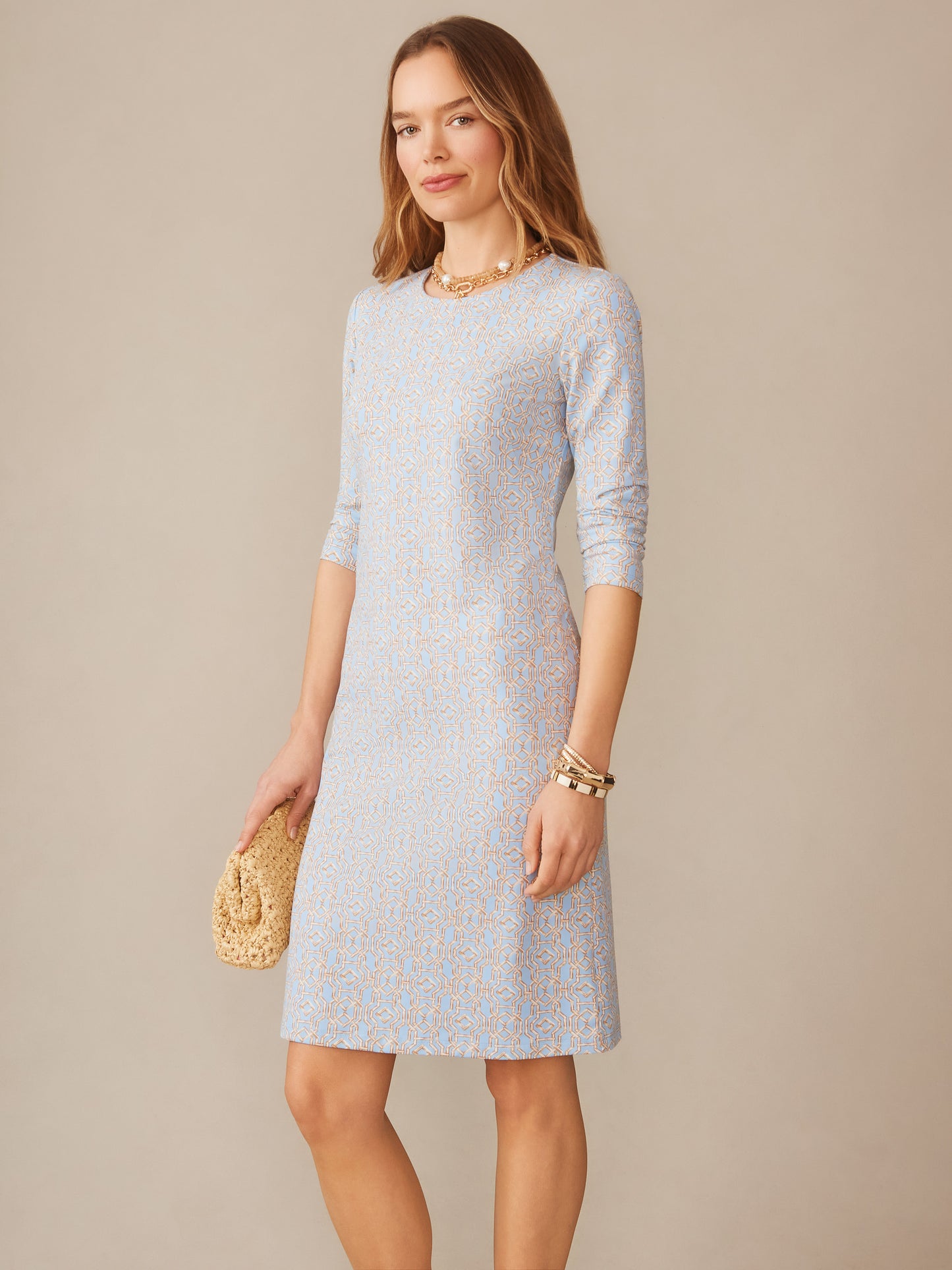 Woman wearing the Sophia Dress in Bamboo Lane by J.McLaughlin, featuring a refined blue and brown pattern dress with three-quarter sleeves, styled with gold bracelets and a woven clutch, embodying effortlessly stylish, enduring designs and innovative fabrics from a destination for defining style. Life, Well-Lived.