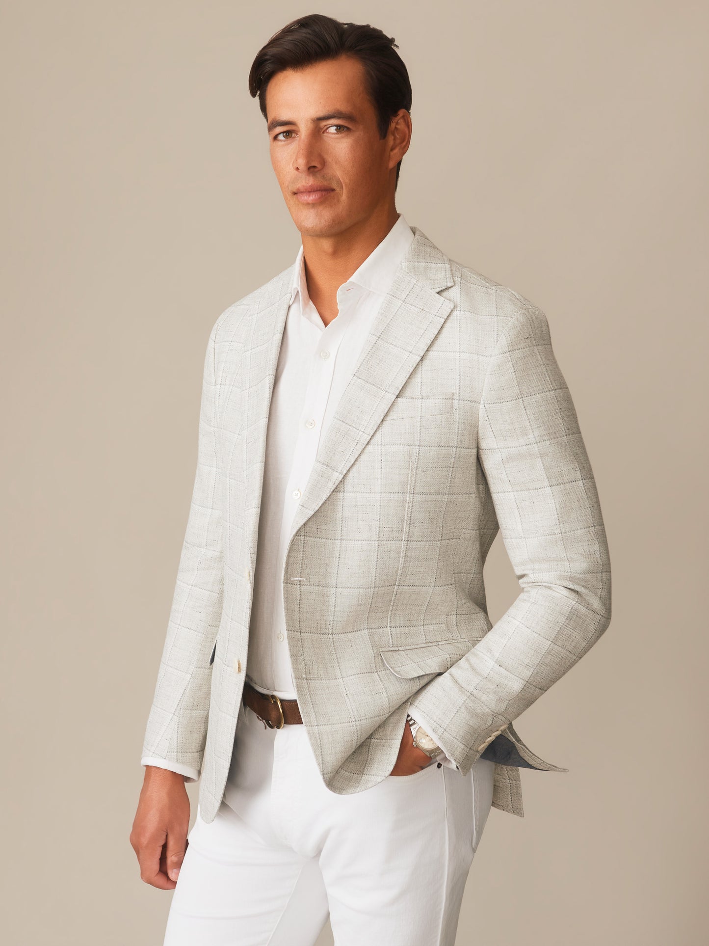 A man wearing a Suffolk Linen Sport Coat in Window Pane by J.McLaughlin, featuring a light gray checked pattern, styled with a white shirt and white pants, embodying effortlessly stylish, enduring designs and innovative fabrics for a legacy of timeless fashion. A destination for defining style. Life, Well-Lived.