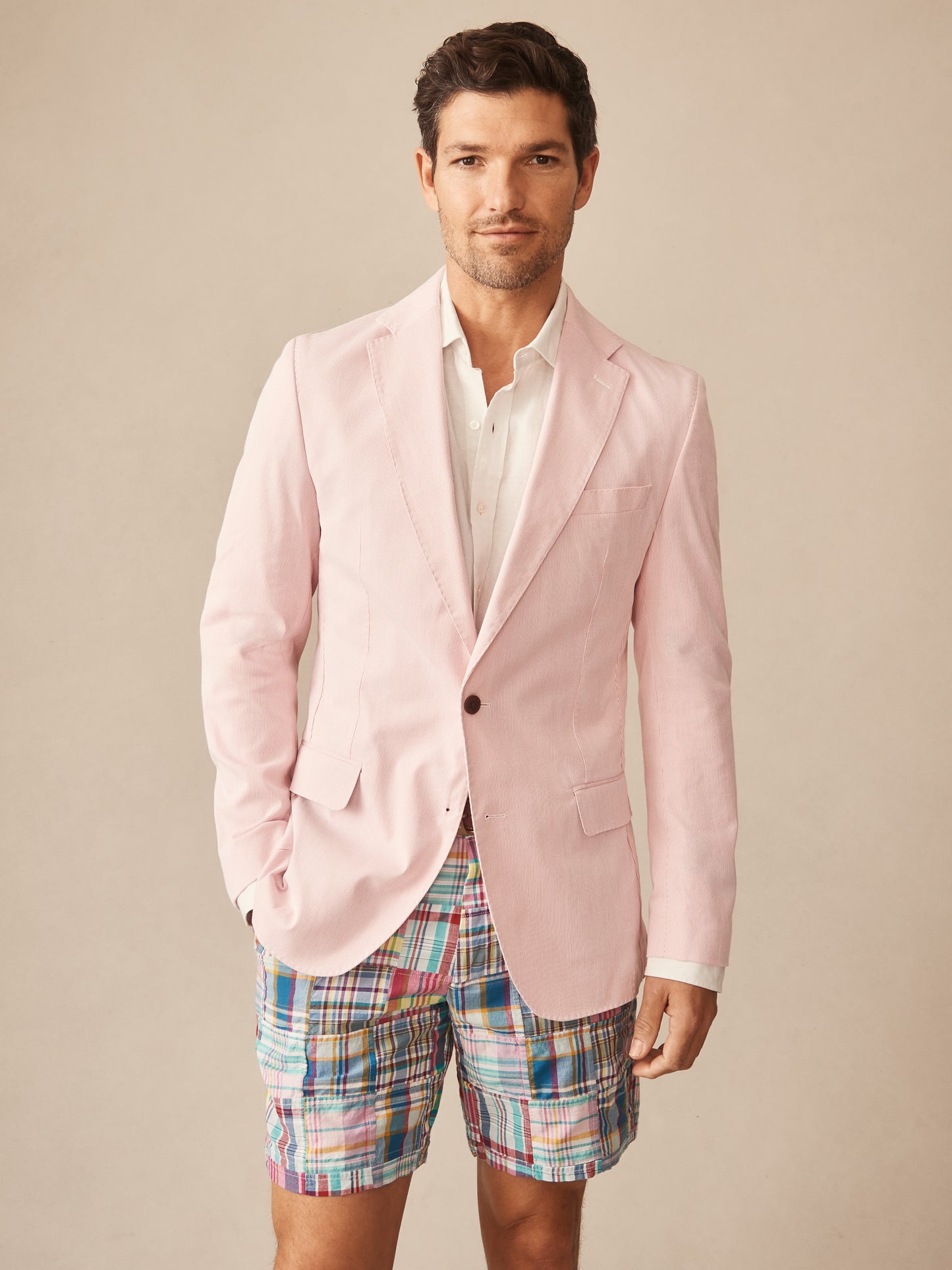 A man wears the Suffolk Sport Coat in Fineline by J.McLaughlin, featuring a soft pink tone paired with colorful plaid shorts, showcasing effortlessly stylish, enduring designs with innovative fabrics in a legacy of timeless fashion. A destination for defining style. Life, Well-Lived.