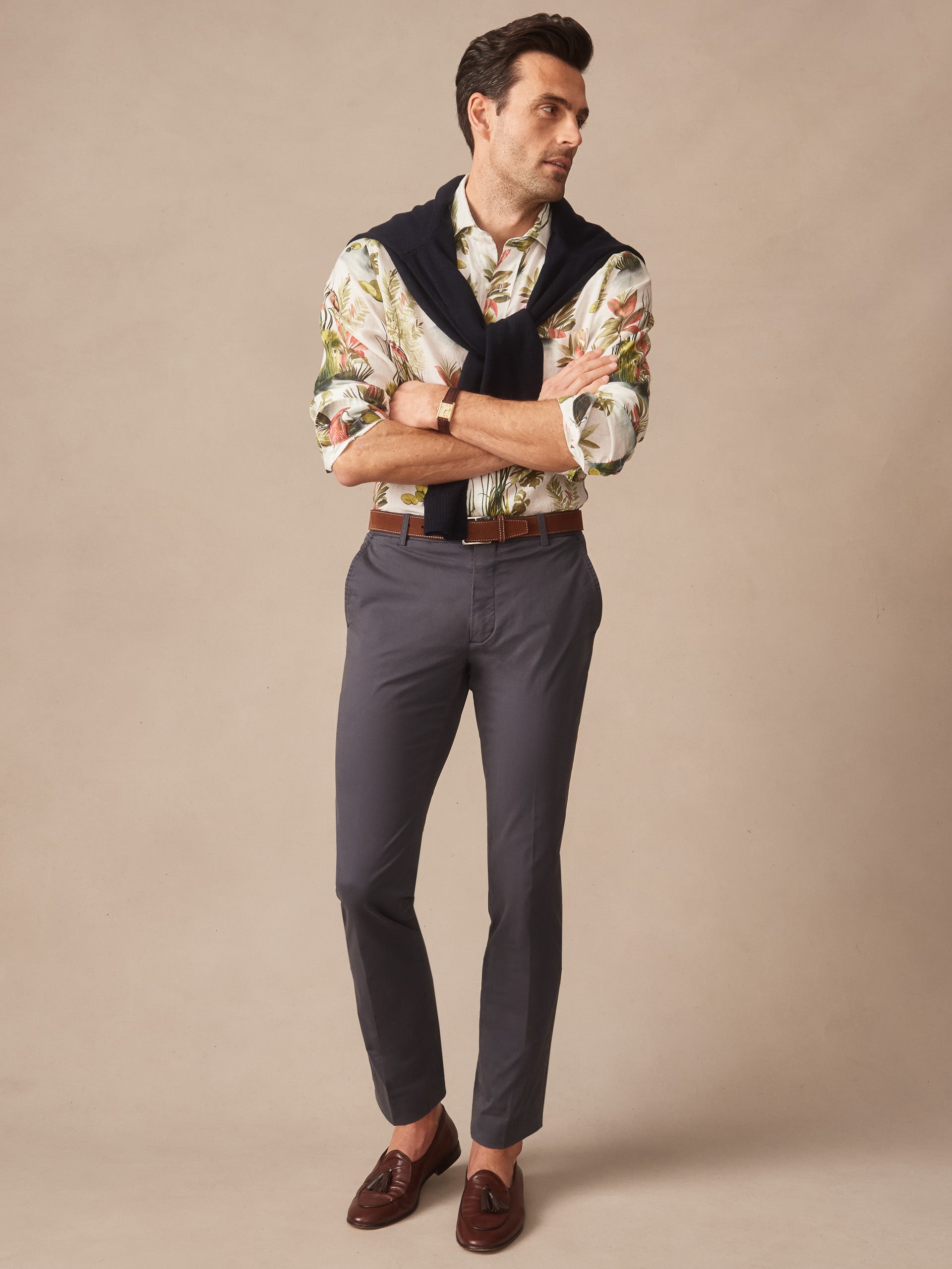 Man wearing J.McLaughlin Madison Pants in washed black paired with brown leather loafers, a floral shirt, and a navy sweater draped over shoulders, showcasing effortlessly stylish, enduring designs with innovative fabrics and a legacy of timeless fashion. A destination for defining style. Life, Well-Lived.