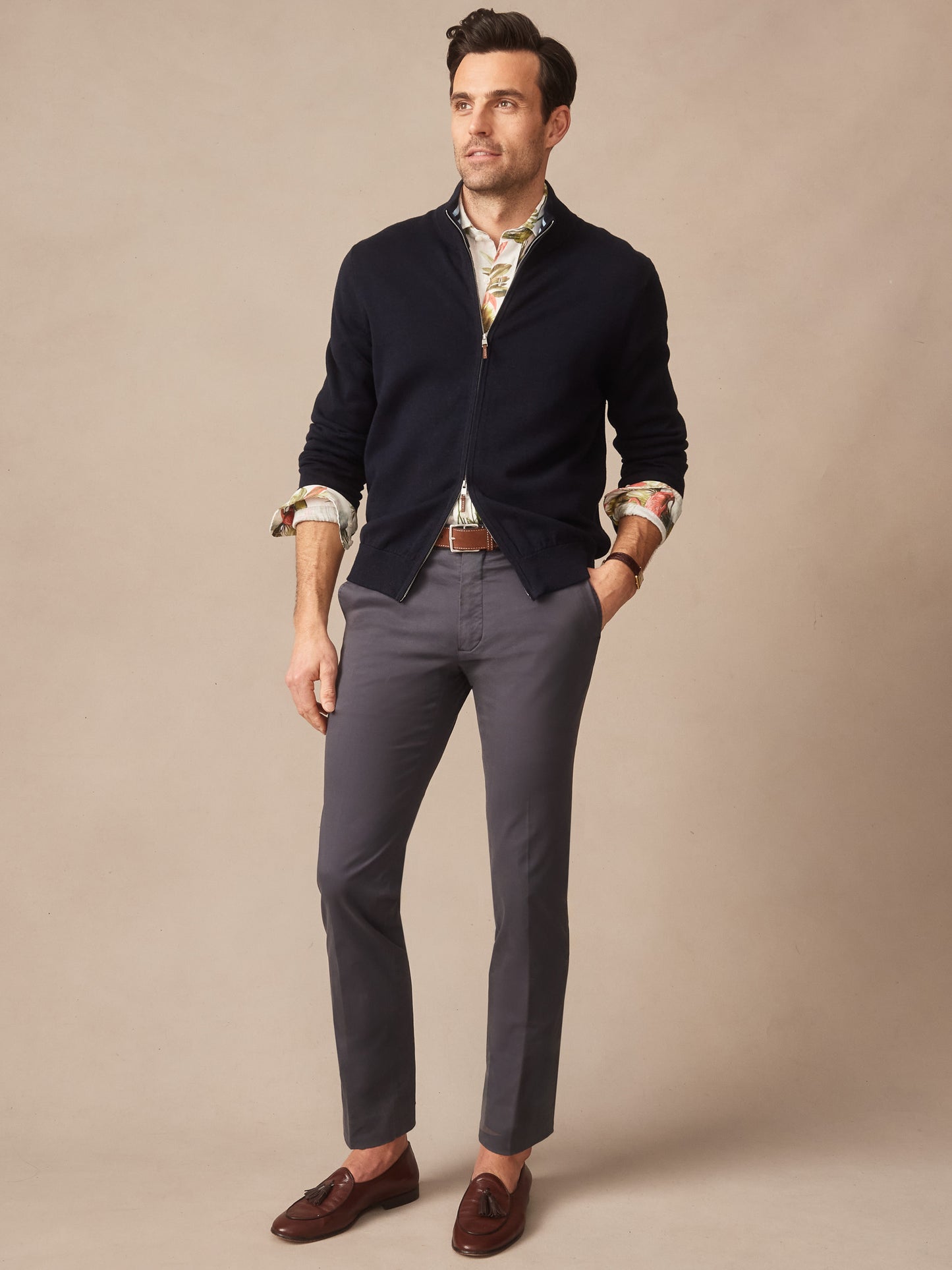 A man models the Orwell Sweater by J.McLaughlin in dark navy, styled over a patterned shirt with grey pants and brown loafers, embodying effortlessly stylish, enduring designs with innovative fabrics and a legacy of timeless fashion. A destination for defining style. Life, Well-Lived.