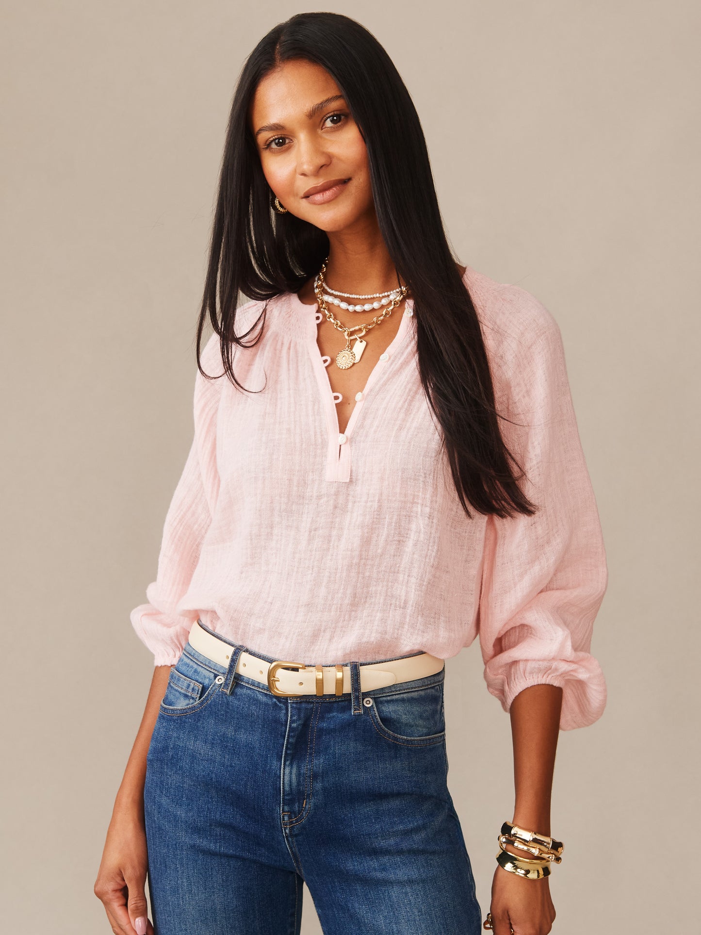 Woman wearing a soft pink Tatiana Linen Blouse by J.McLaughlin paired with blue jeans and gold accessories, showcasing effortlessly stylish, enduring designs with innovative fabrics.