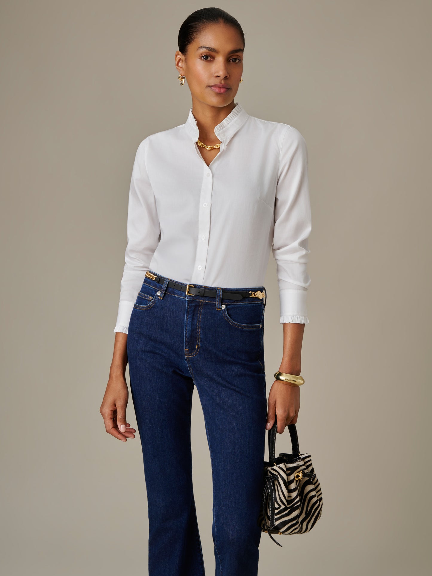 Woman wearing a white Tess Ruffle Collar Shirt by J.McLaughlin paired with dark blue jeans, accessorized with gold jewelry and a small zebra print handbag, showcasing timeless style.