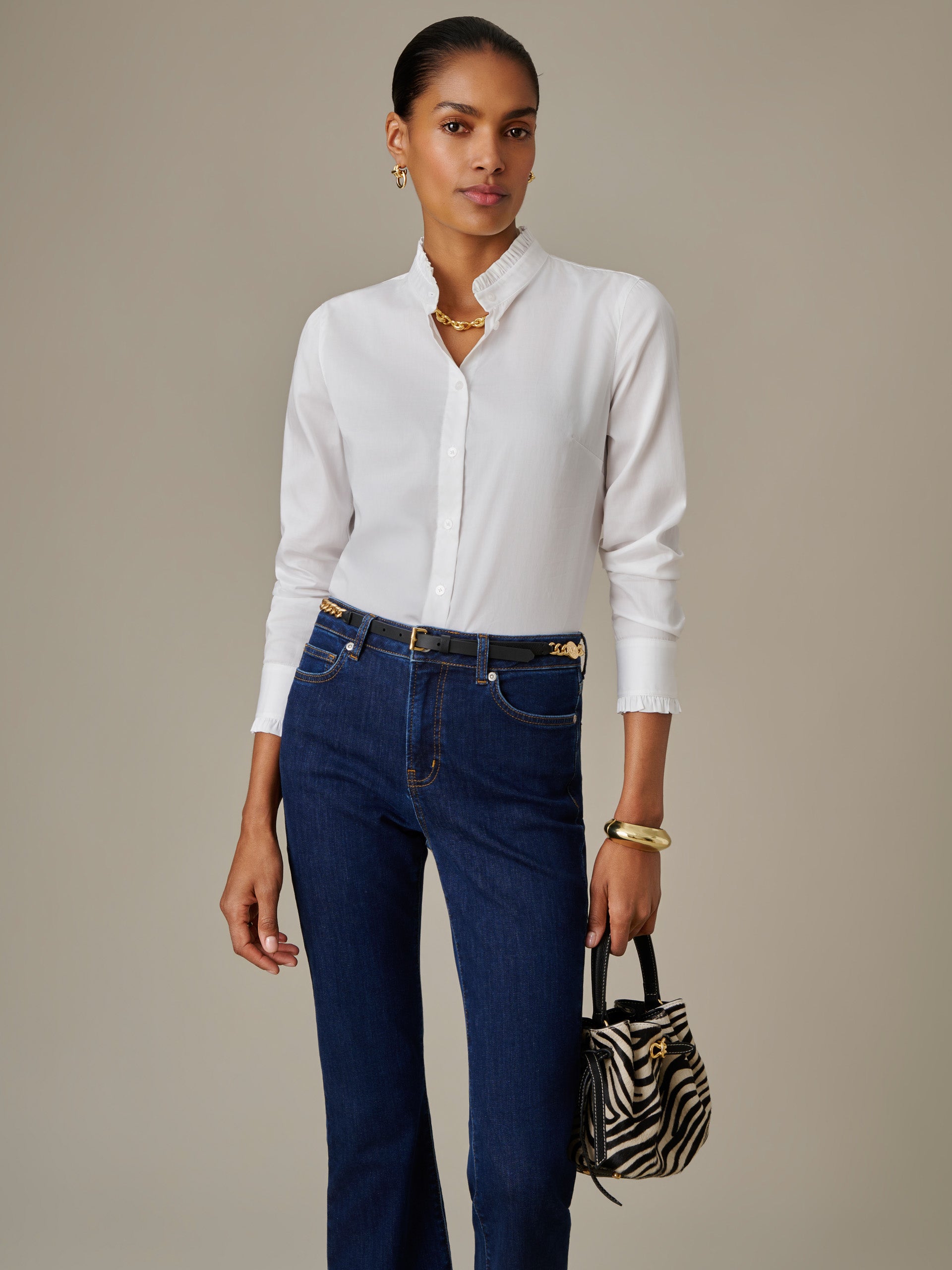 Woman wearing a white Tess Ruffle Collar Shirt by J.McLaughlin paired with dark blue jeans, accessorized with gold jewelry and a small zebra print handbag, showcasing timeless style.