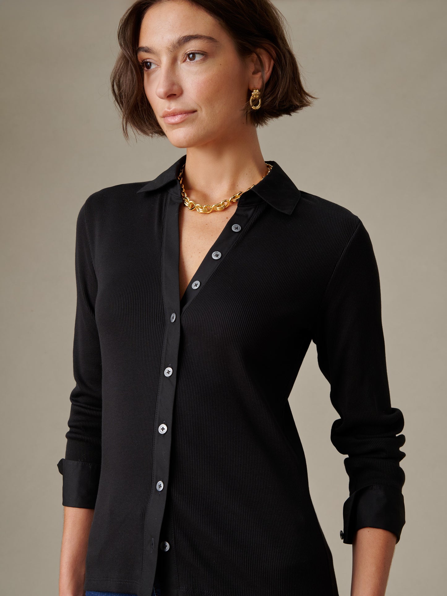 Woman wearing a black Theodora Shirt by J.McLaughlin, styled with gold chain necklace and hoop earrings, showcasing effortlessly stylish and enduring designs.