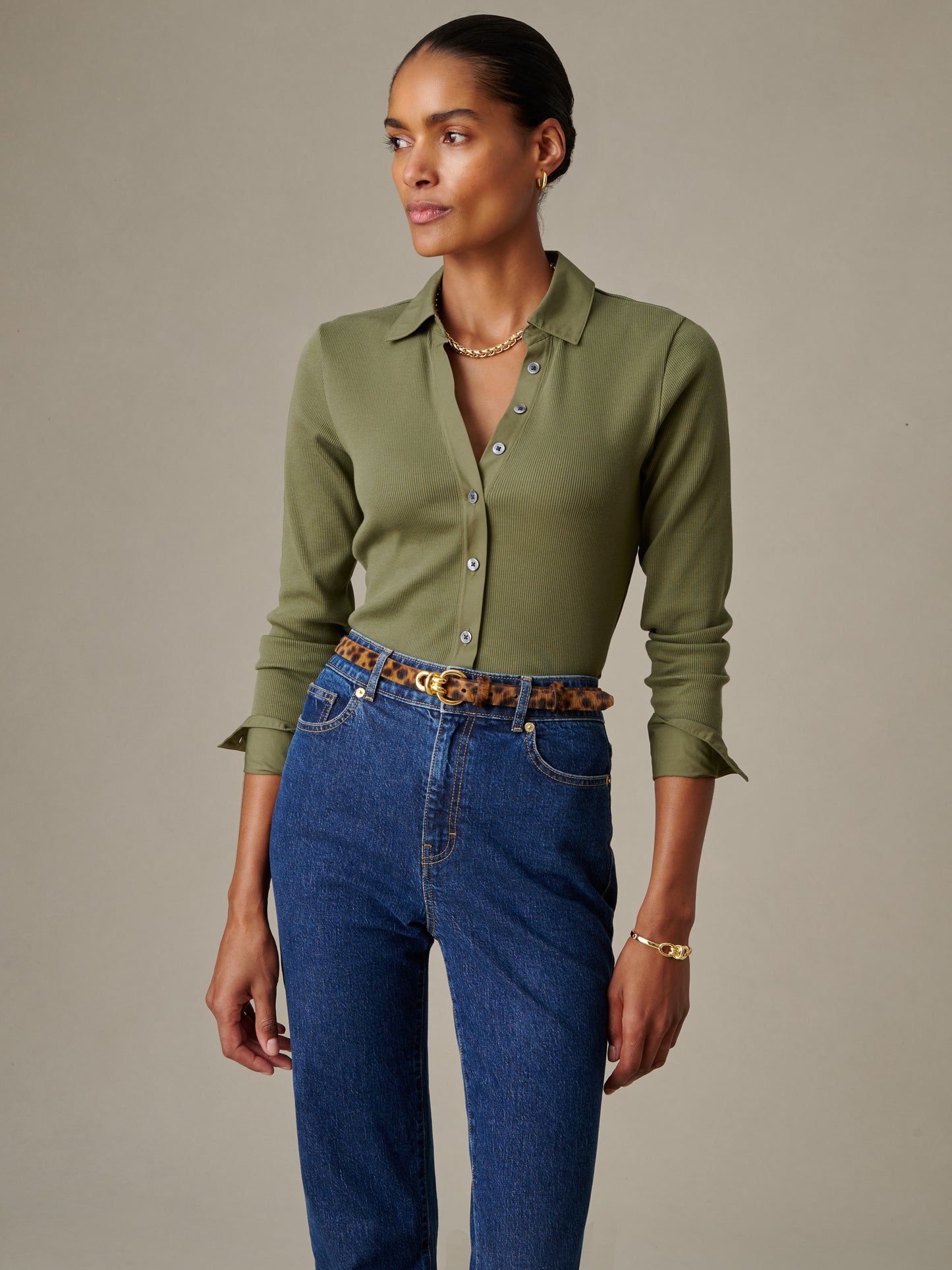 A model wearing the olive green Theodora Shirt by J.McLaughlin paired with high-waisted jeans and a leopard print belt, showcasing effortlessly stylish, enduring designs.