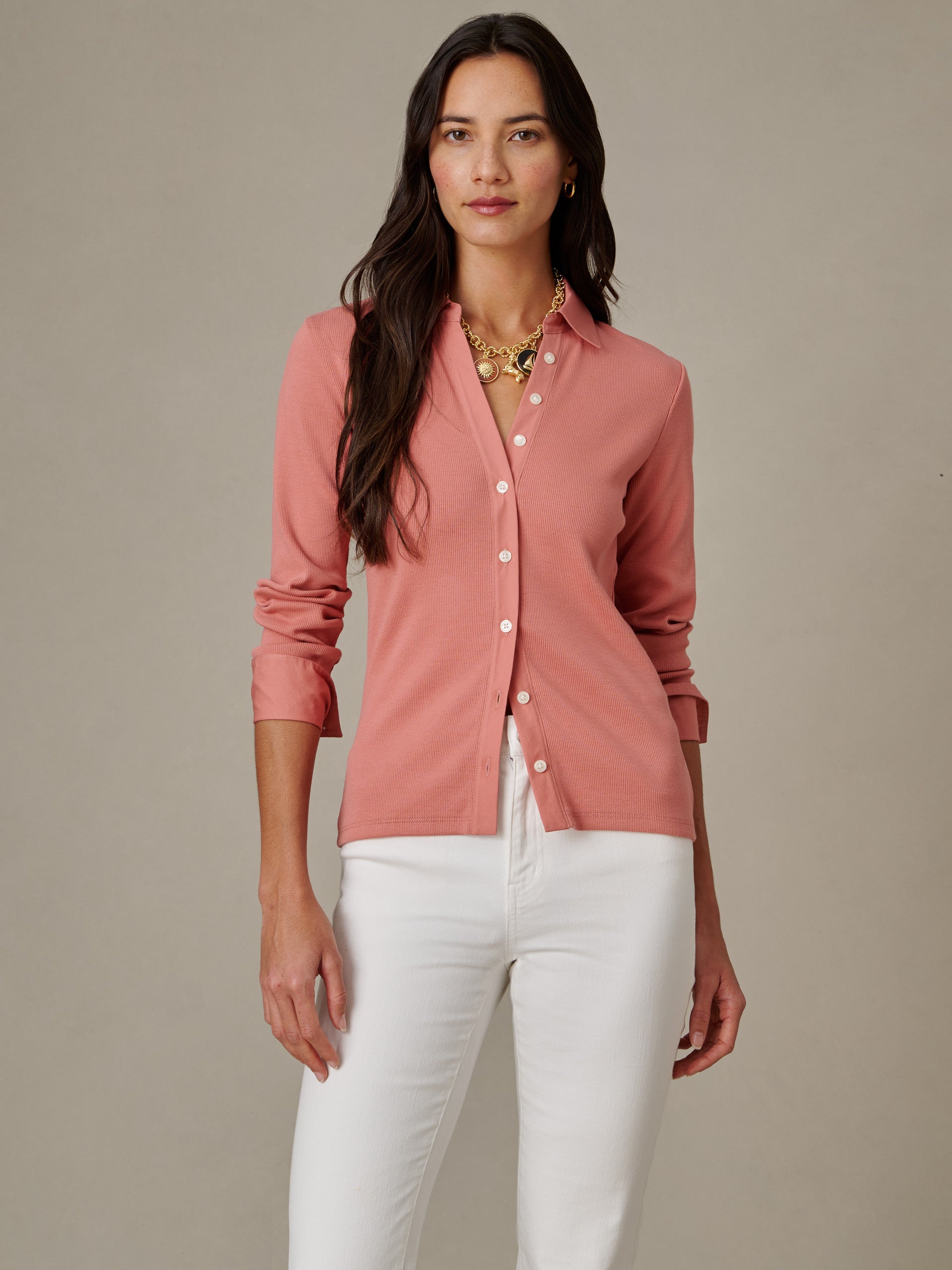Woman wearing a long-sleeve, button-down Theodora Shirt in soft pink paired with white pants, styled with layered gold necklaces, showcasing J.McLaughlin's enduring and effortlessly stylish design.