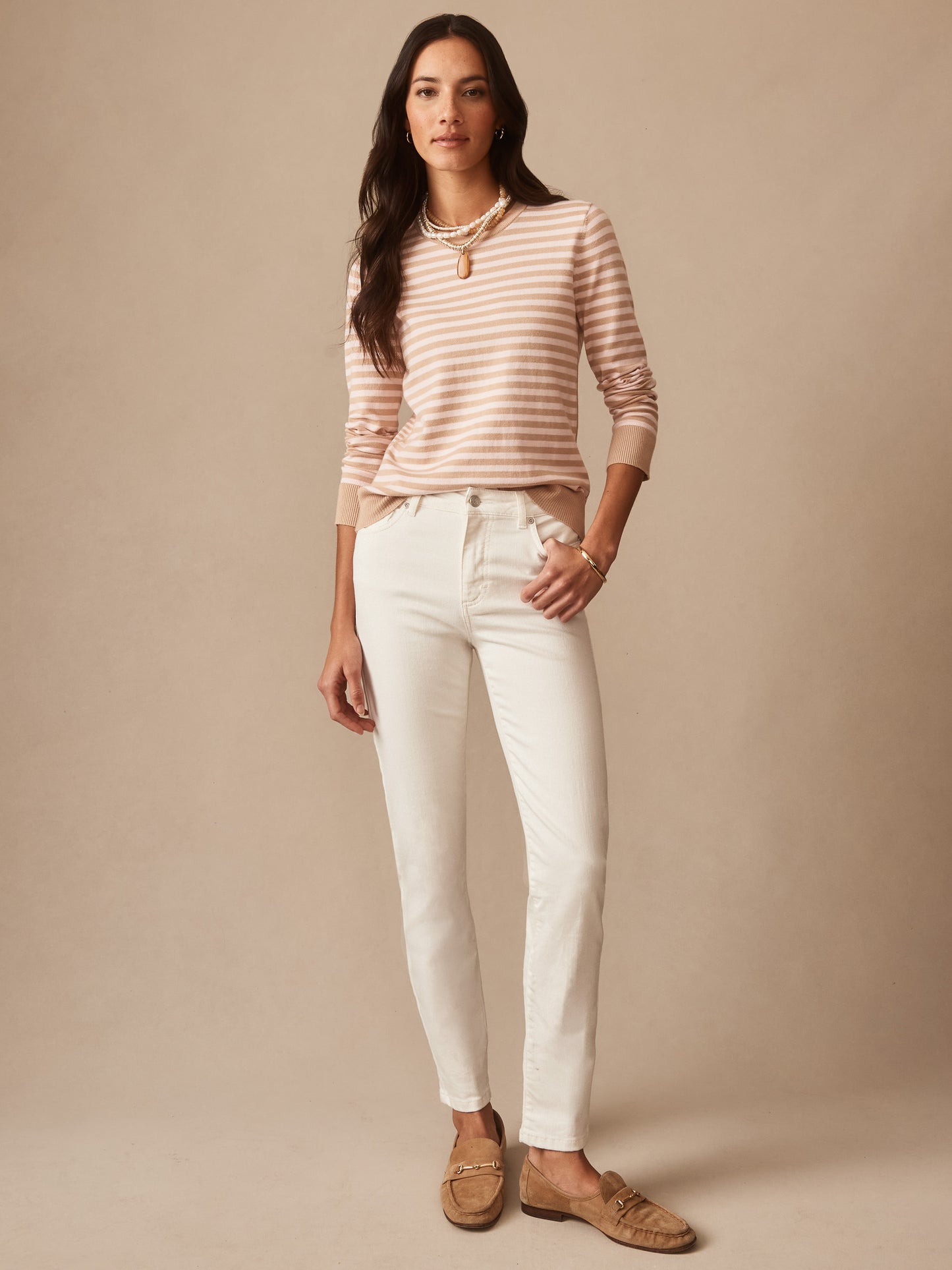 Woman wearing Watson Jeans by J.McLaughlin in a natural white shade paired with a beige and pink striped long-sleeve top and tan loafers, styled with layered necklaces.