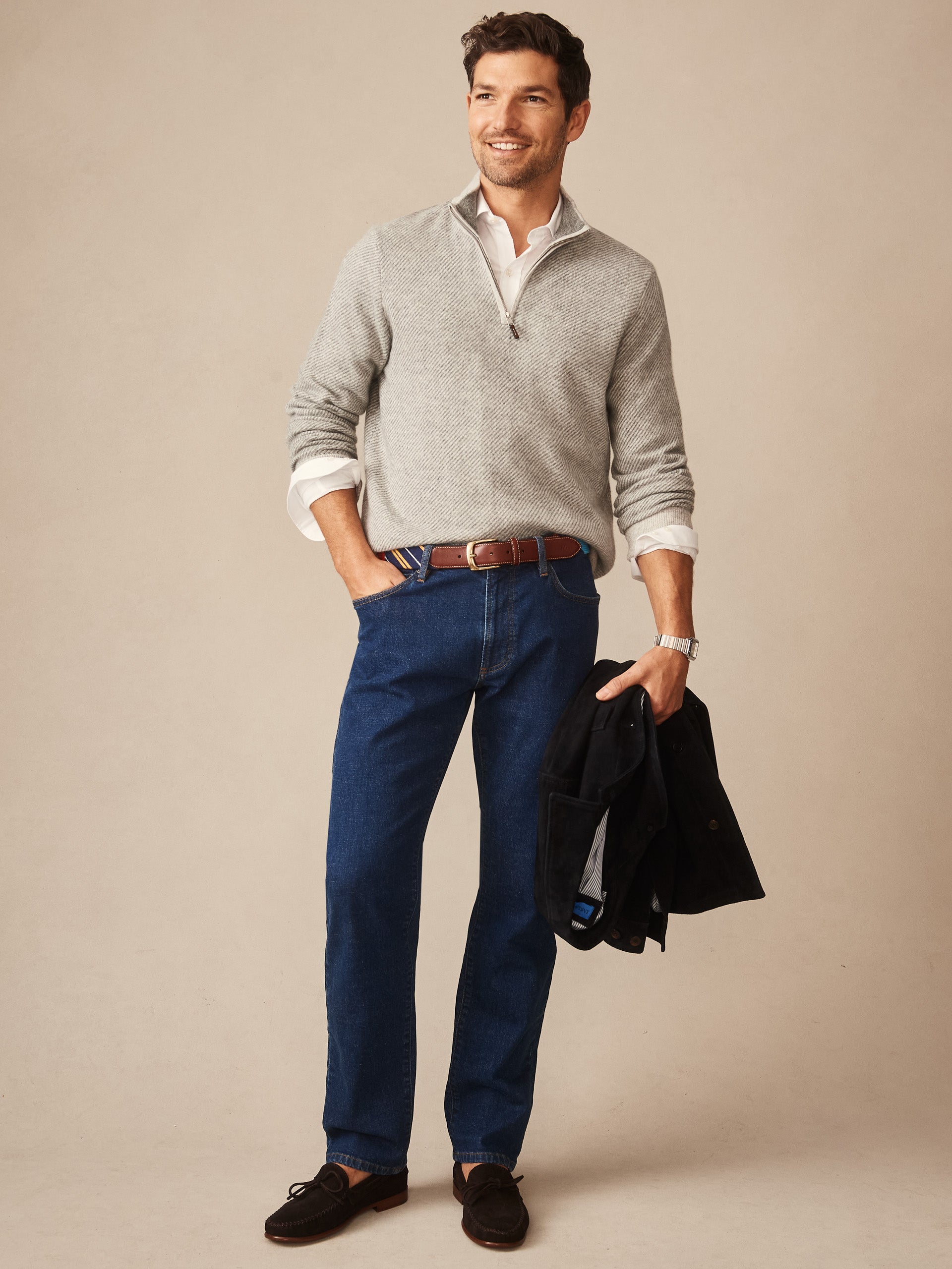Man wearing a gray Twain Cashmere 1/4 Zip in Jacquard sweater layered over a white shirt, paired with dark blue jeans and brown belt, holding a black jacket, embodying J.McLaughlin's effortlessly stylish, enduring designs and innovative fabrics. A destination for defining style. Life, Well-Lived.