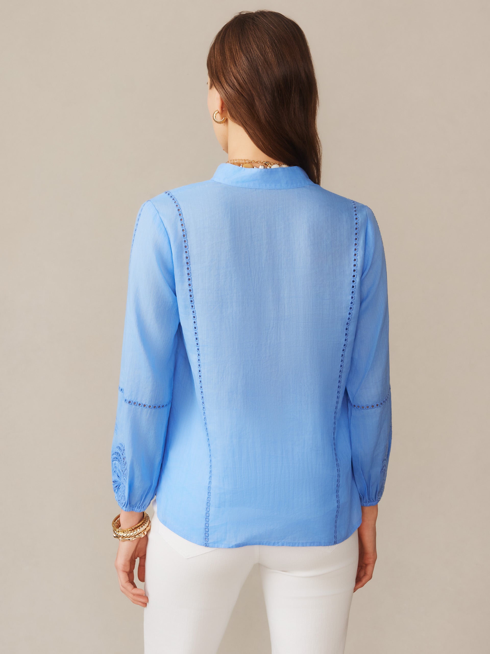 A woman wearing a cornflower blue Varsha Blouse by J.McLaughlin, featuring delicate stitching details on the back and sleeves, embodying effortlessly stylish and enduring designs.