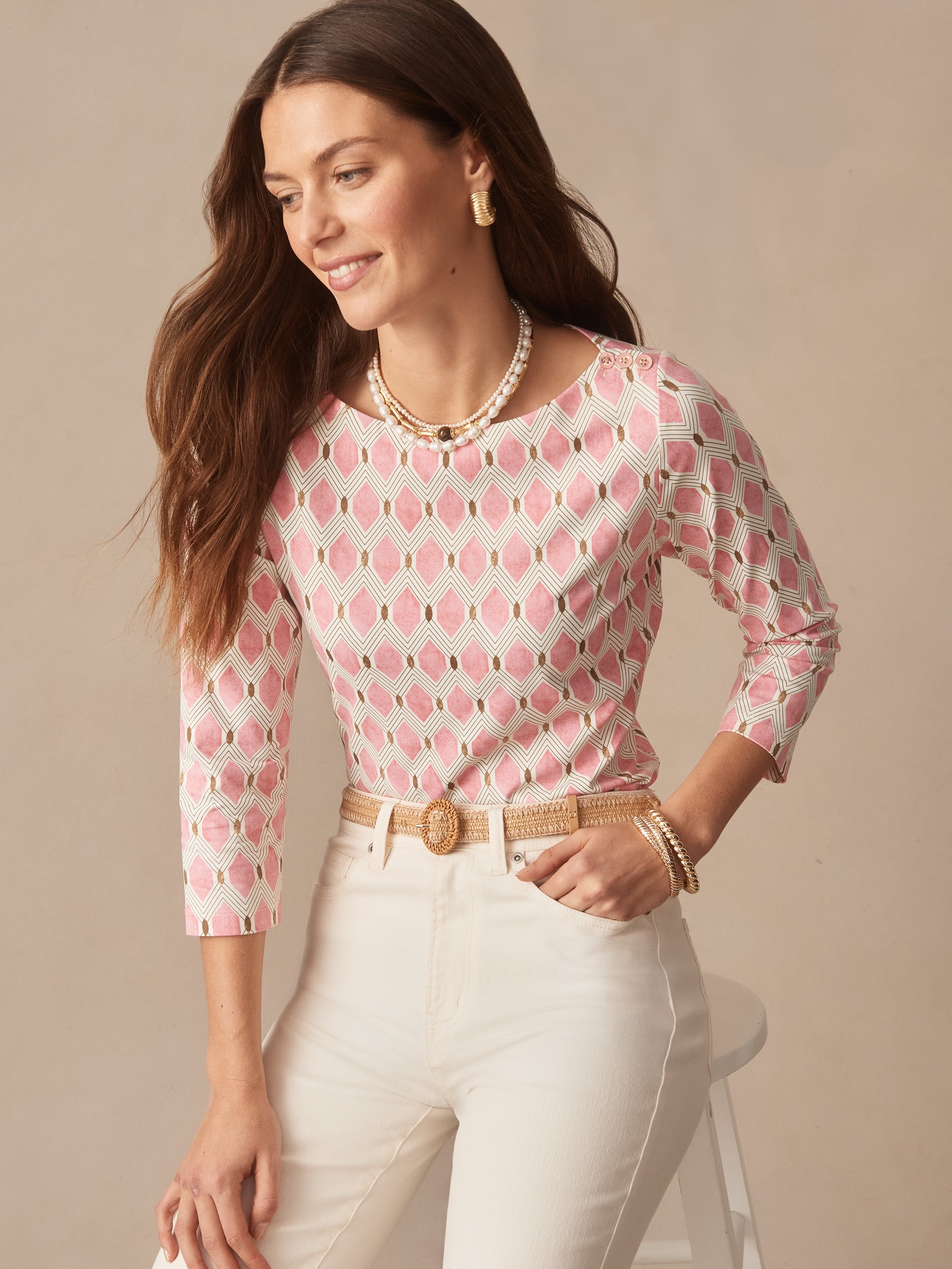 A woman wearing the Wavesong Tee in Mini Beekeeper by J.McLaughlin, featuring a pink and tan patterned top paired with white pants and gold accessories, showcasing effortlessly stylish, enduring designs and innovative fabrics. A destination for defining style. Life, Well-Lived.