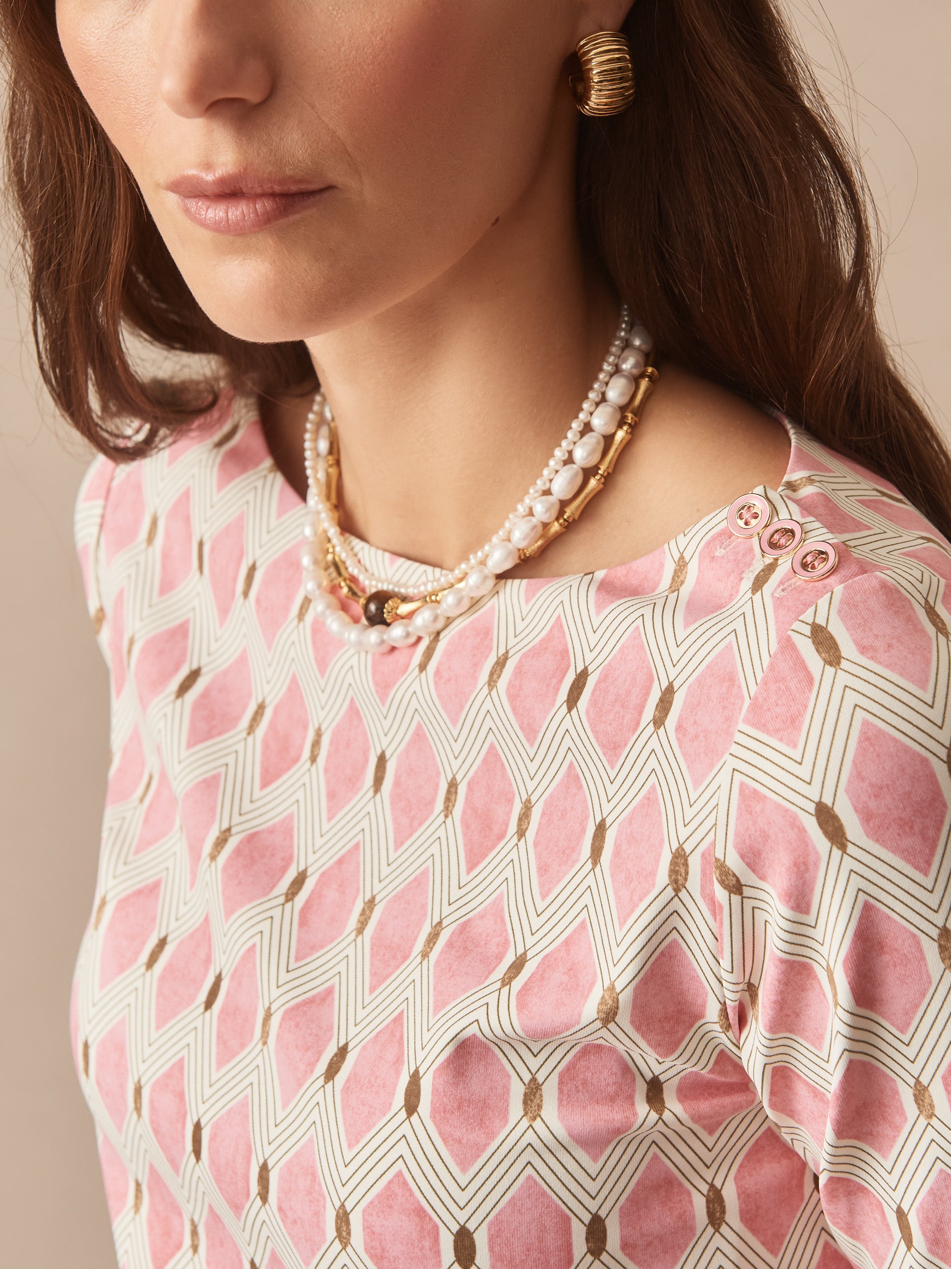 Close-up of a woman wearing the Wavesong Tee in Mini Beekeeper by J.McLaughlin, featuring a pink geometric pattern, gold-tone buttons on the shoulder, and layered pearl necklaces, showcasing effortlessly stylish design and enduring appeal from a destination for defining style. Life, Well-Lived.