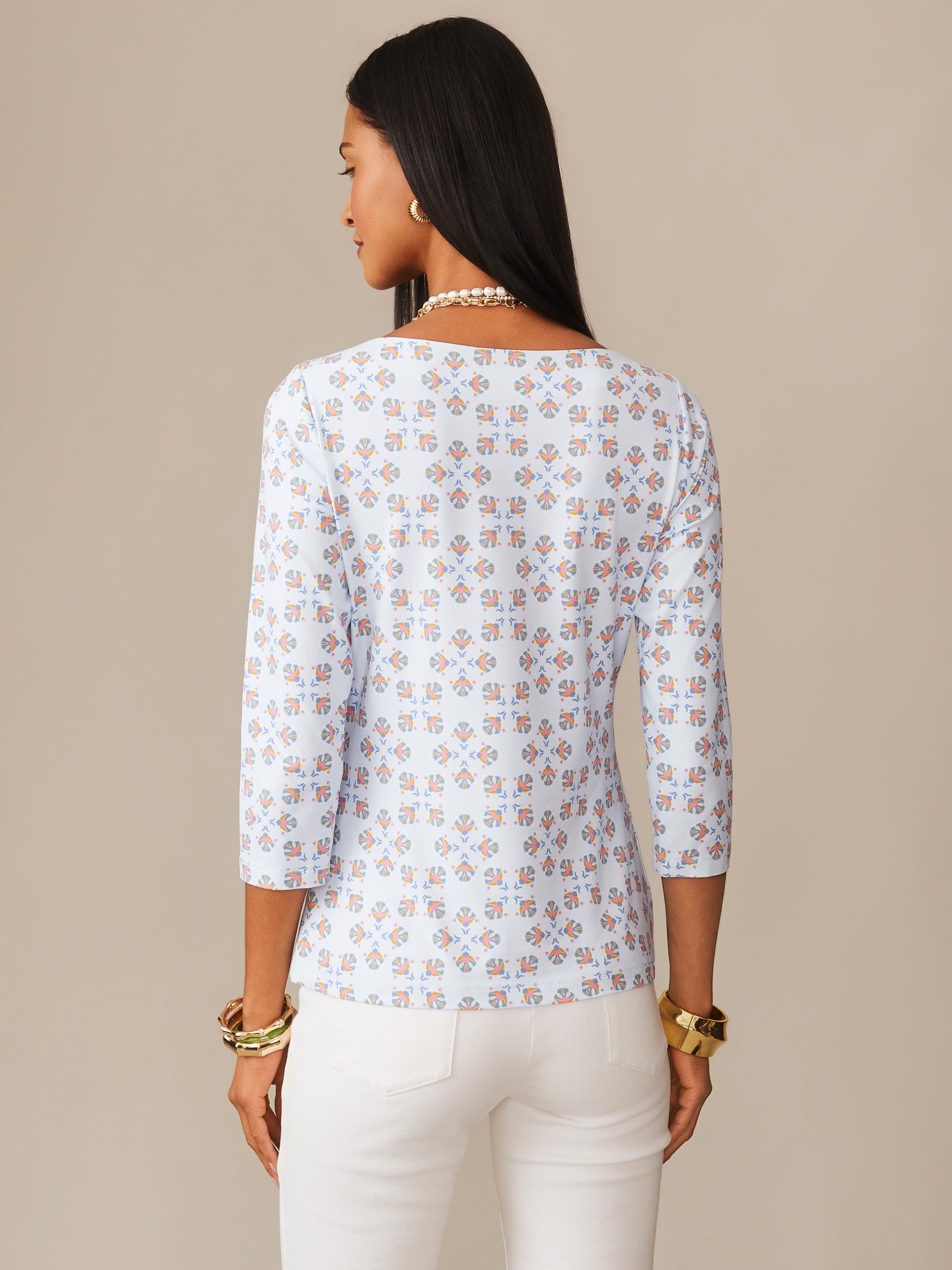 Woman wearing a light blue Wavesong Tee in Mythic Tile by J.McLaughlin, featuring an all-over geometric pattern, styled with white pants and gold accessories, showcasing effortlessly stylish, enduring designs and innovative fabrics in a legacy of timeless fashion. A destination for defining style. Life, Well-Lived.