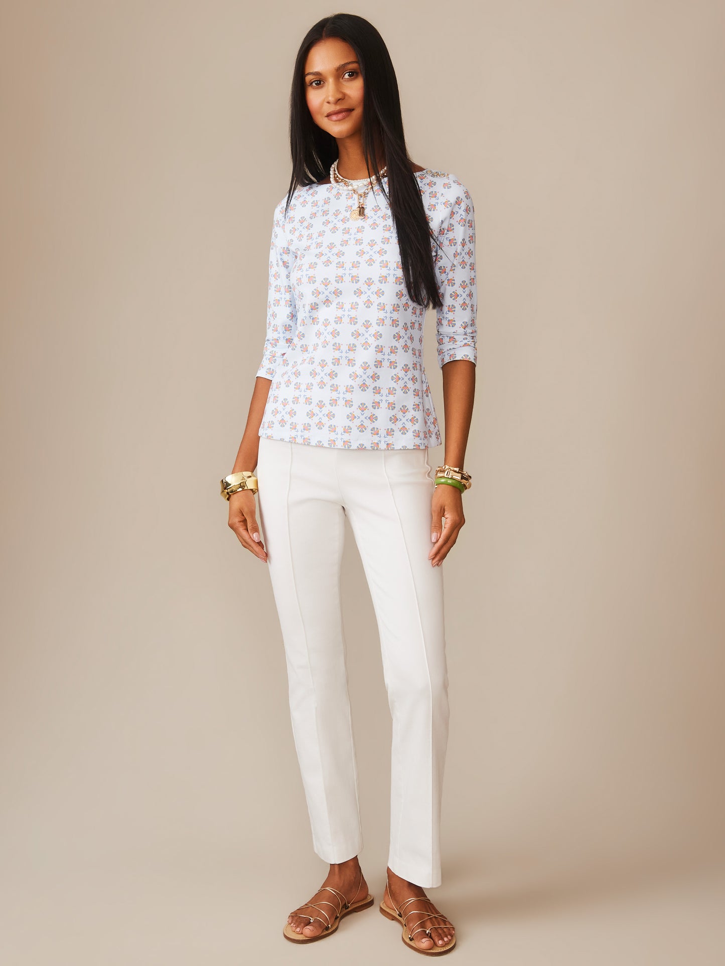 A confident woman models the Wavesong Tee in Mythic Tile by J.McLaughlin, featuring a light blue patterned top paired with white pants and gold accessories, embodying effortlessly stylish, enduring designs crafted with innovative fabrics—a destination for defining style. Life, Well-Lived.