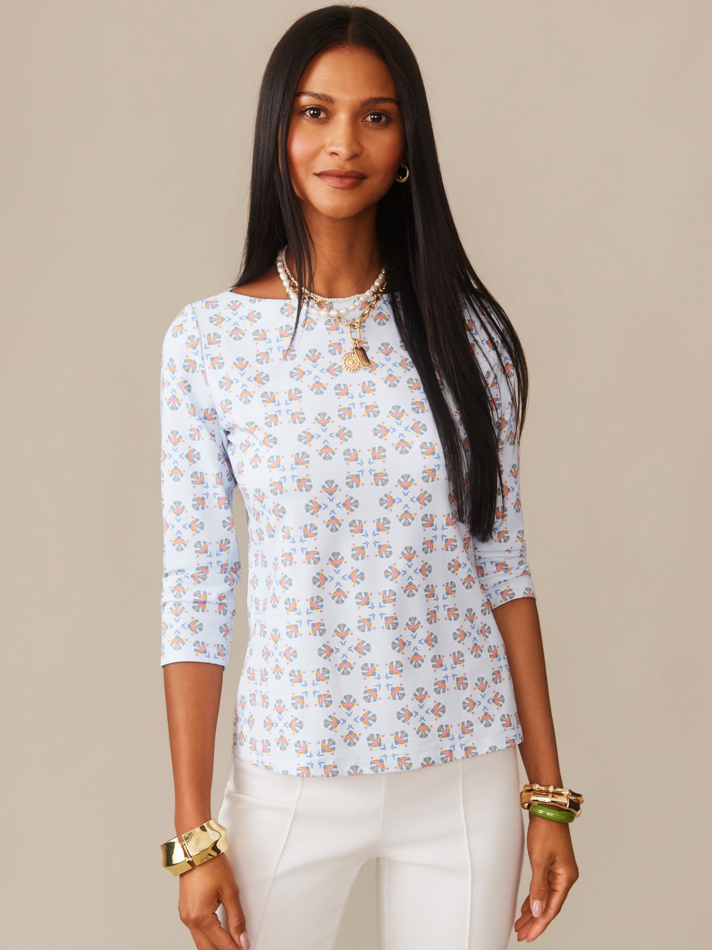 Woman wearing a light blue Wavesong Tee in Mythic Tile patterned top by J.McLaughlin, styled with white pants and gold jewelry, embodying effortlessly stylish, enduring designs and a legacy of timeless fashion.