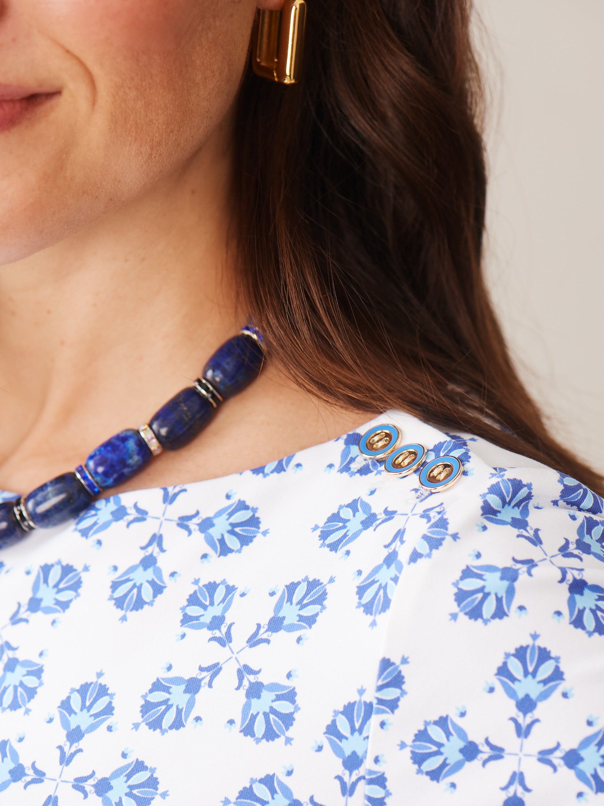 Close-up of a woman wearing the Wavesong Tee in Mythic Tile by J.McLaughlin, featuring a white fabric with a blue floral pattern, accented by gold buttons and paired with a bold blue beaded necklace, showcasing effortlessly stylish, enduring designs with innovative fabrics from a destination for defining style. Life, Well-Lived.